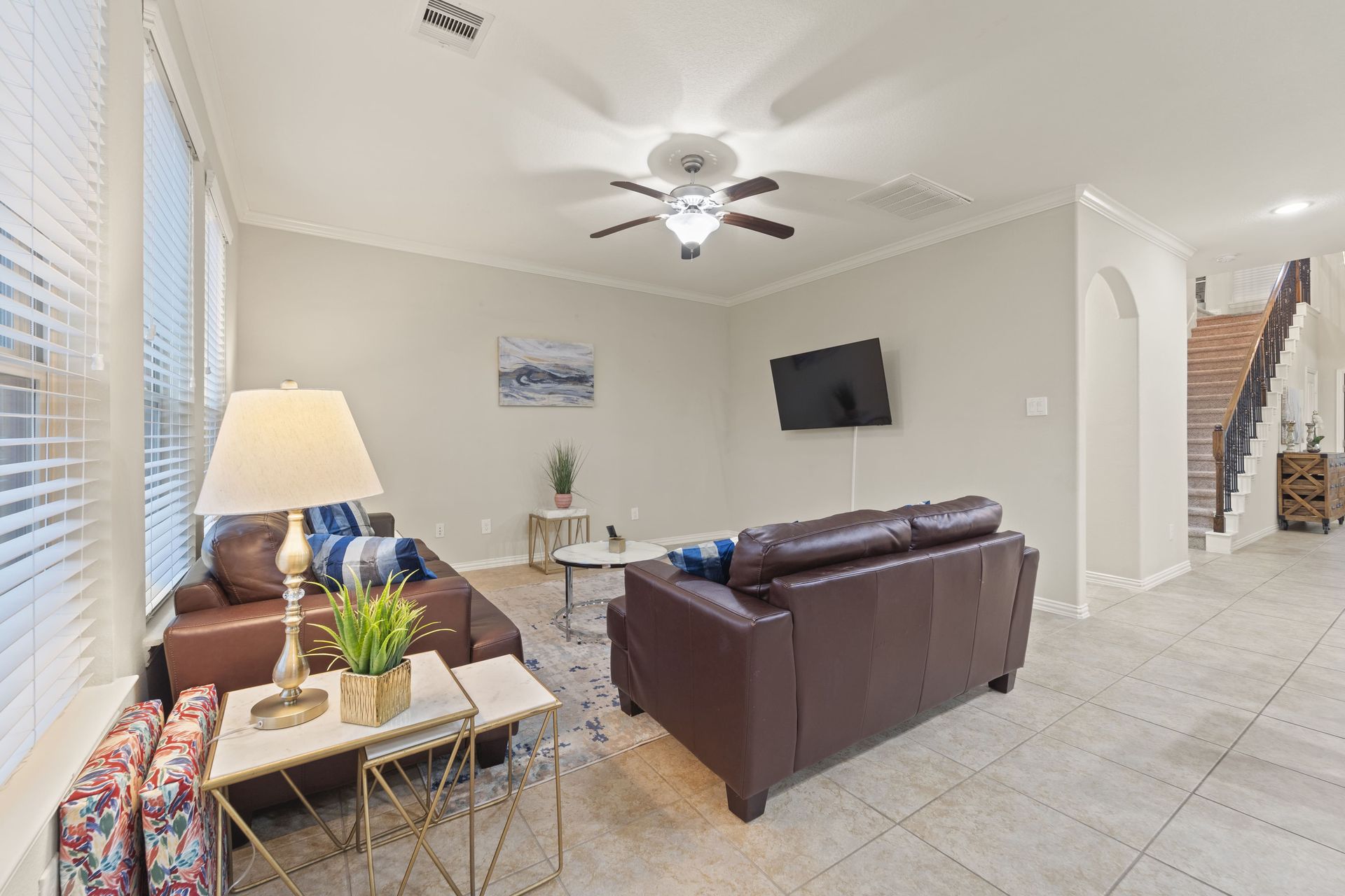 A living room with brown leather sofas, a mounted TV, and a staircase. Natural light streams through the blinds.