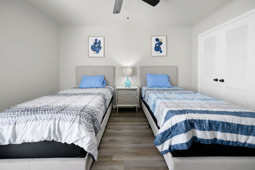 Two twin beds with blue and white bedding in a bedroom with light gray walls and wood-look flooring.