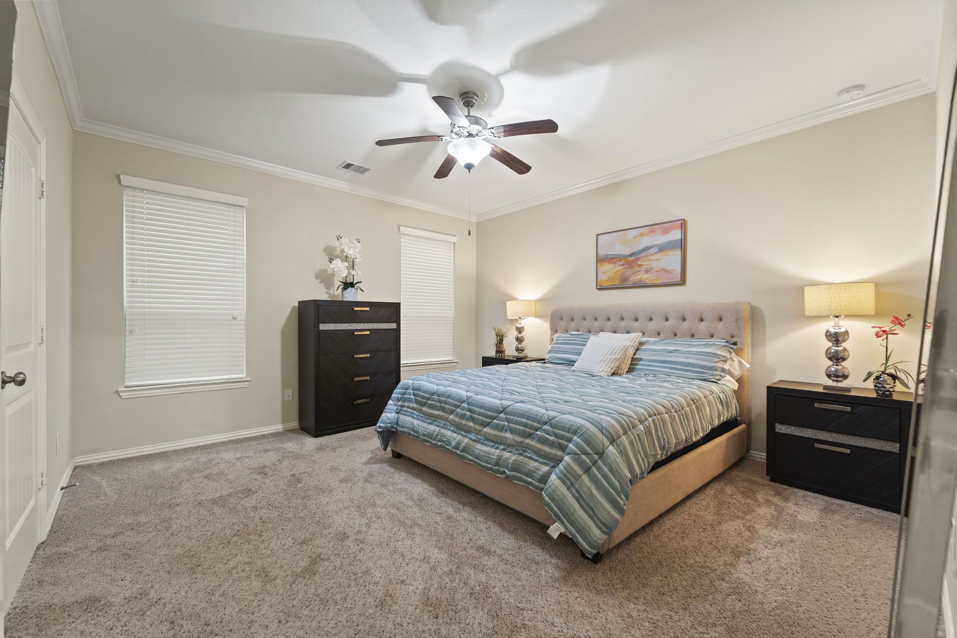 Bedroom with a bed, two nightstands, a dresser, and two windows with blinds. The room is beige and carpeted.
