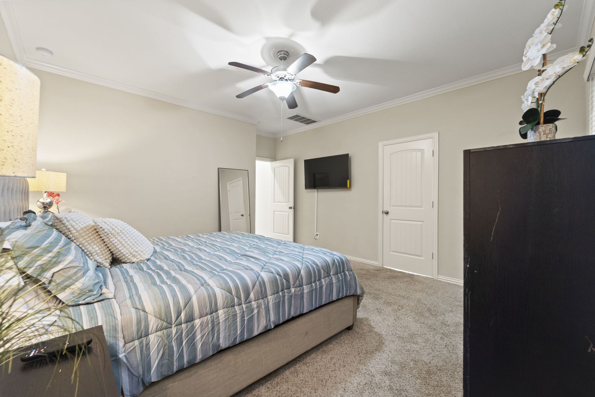 Bedroom with bed, dresser, TV, two doors, and a ceiling fan. Neutral walls, blue and white bedspread, and carpet flooring.