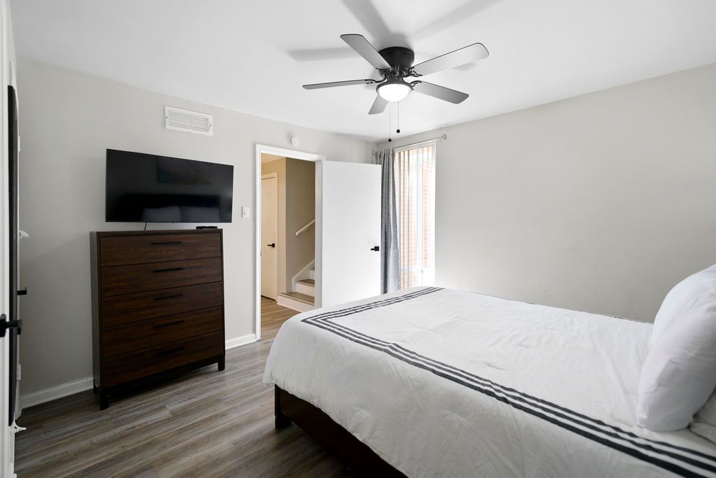 Bedroom with a bed, dresser with a TV, ceiling fan, and an open doorway to a hallway. White bedding and light gray walls.