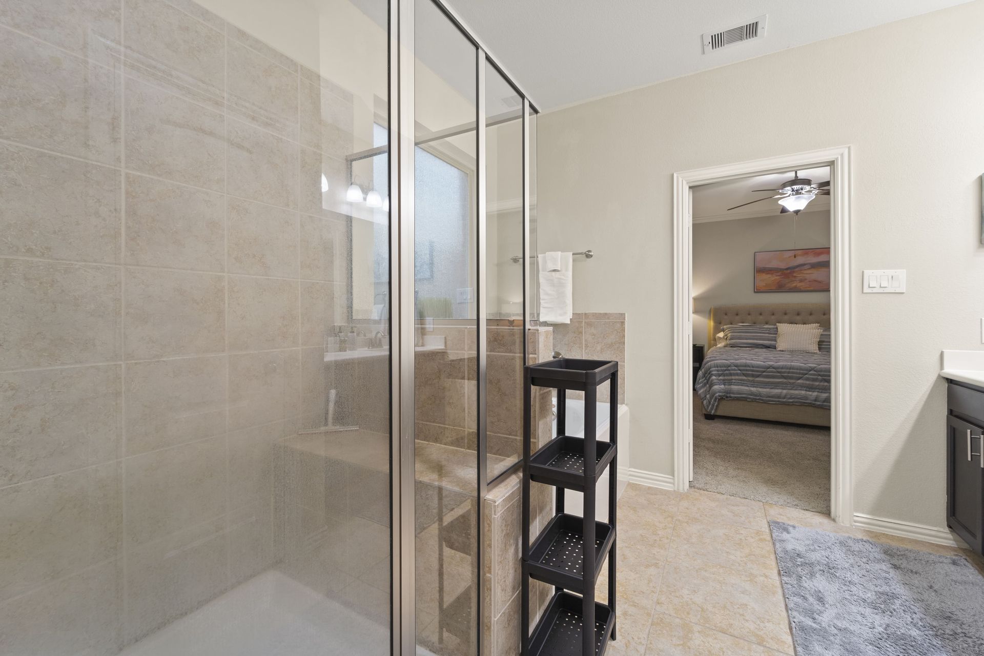 Bathroom with glass shower, gray tiled walls, and a view into a bedroom with a bed.