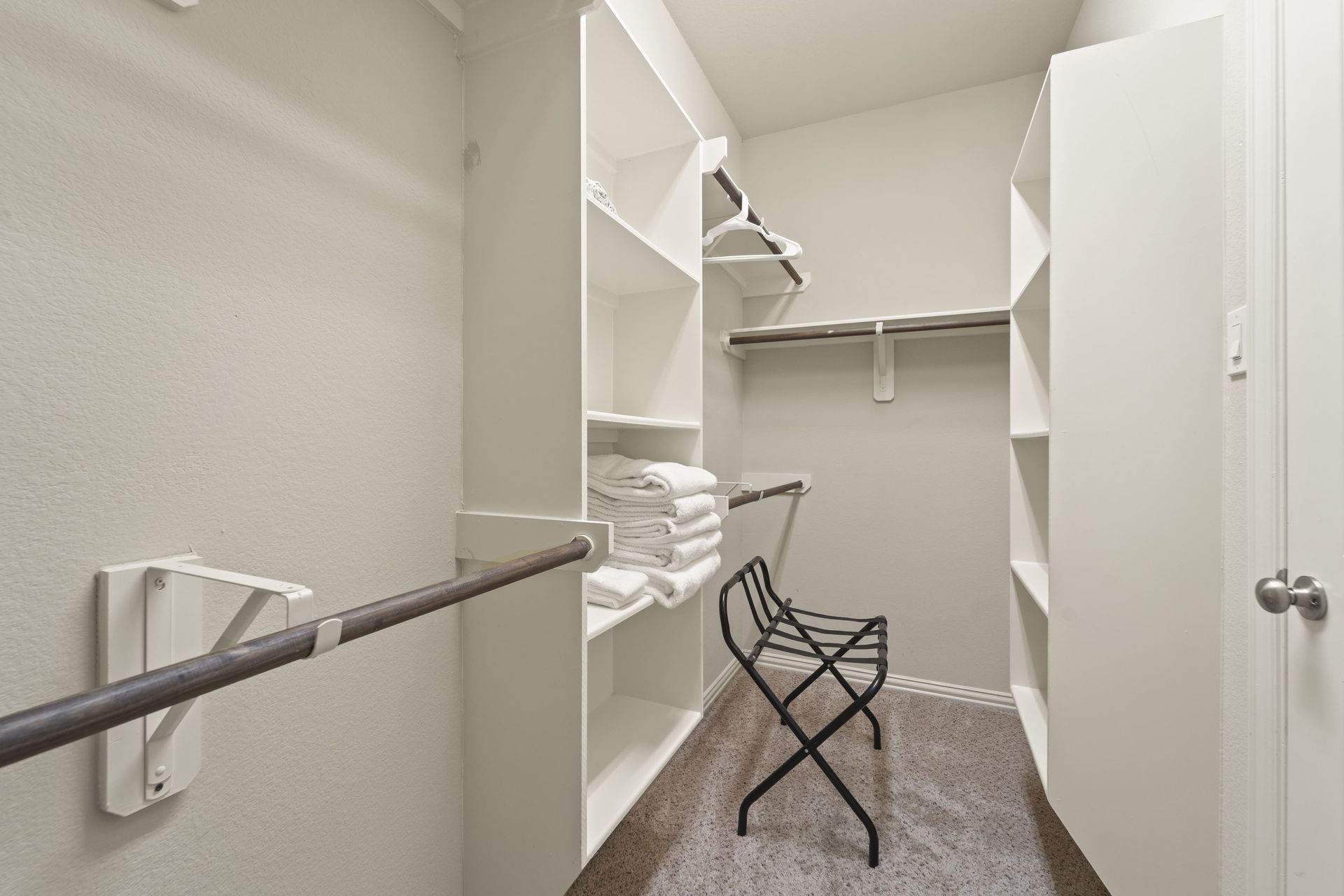 Walk-in closet with white shelving, a dark-colored clothes rod, and a black folding chair. The walls are light-colored.