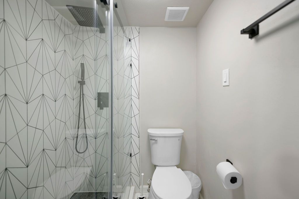 Small, modern bathroom with a white toilet, geometric tiled shower, and gray walls. A black towel bar and toilet paper roll are visible.
