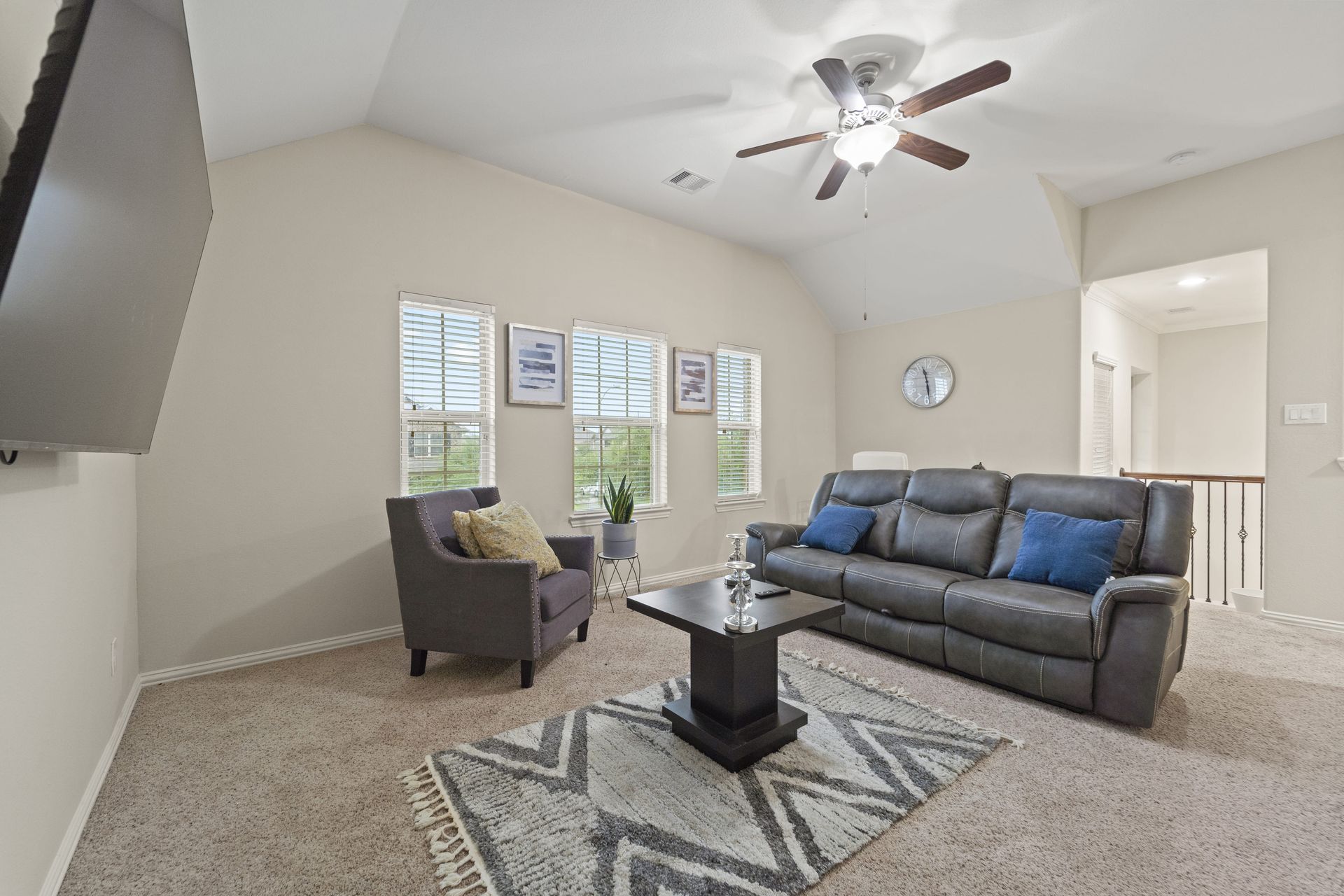 A living room with a gray sofa, armchair, and coffee table on a patterned rug. Three windows are on the back wall.