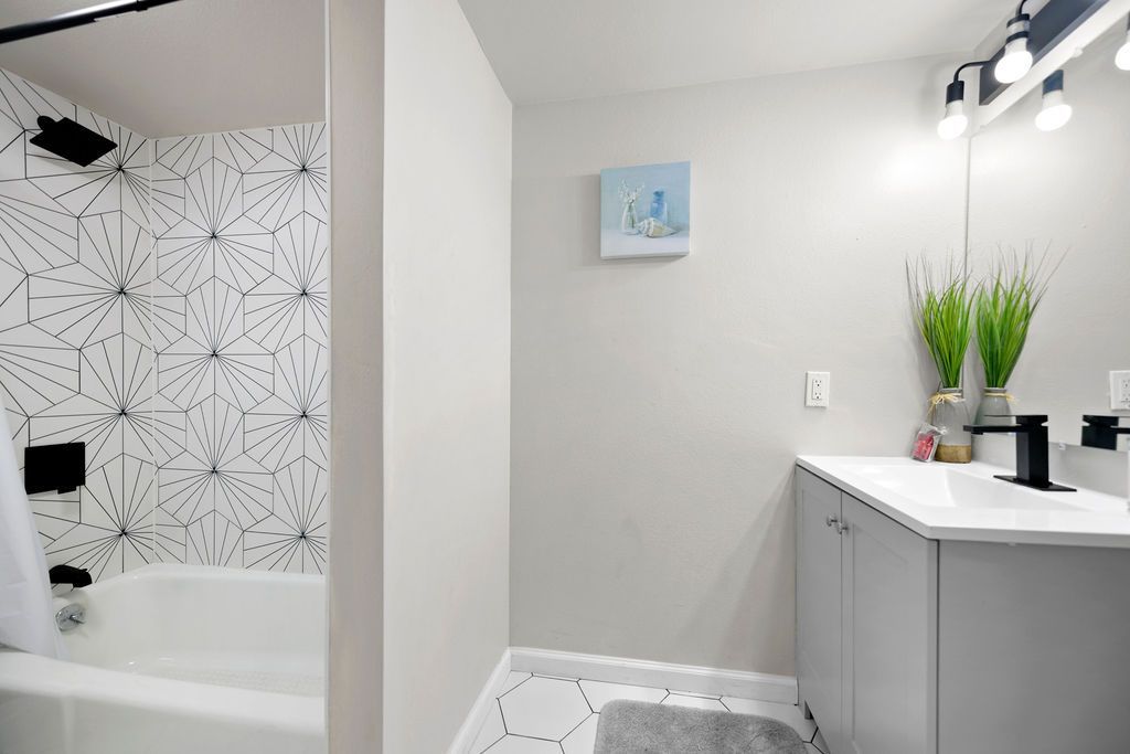 A modern bathroom with a white bathtub, geometric tiled shower wall, and a gray vanity with plants and artwork.