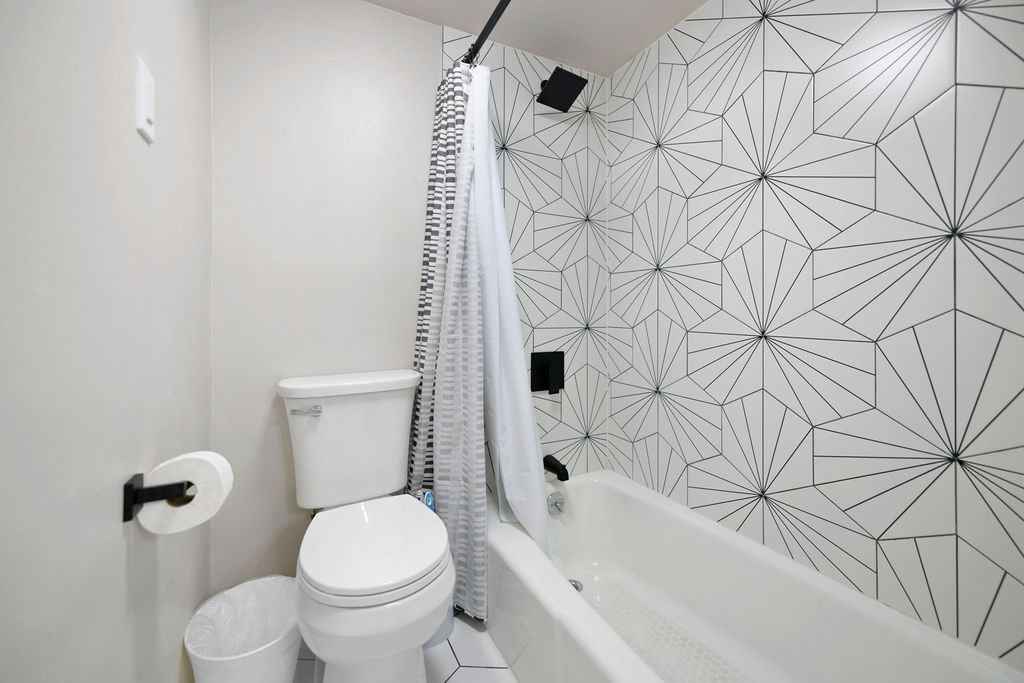 Small bathroom with white and geometric patterned tile, toilet, and a bathtub with a shower curtain.
