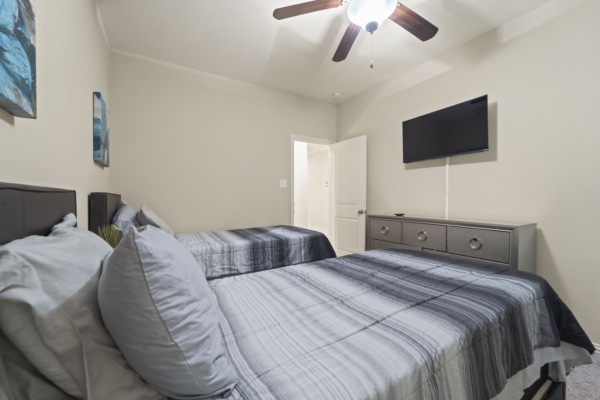 Bedroom with two beds, gray bedding, and a mounted TV. Beige walls, ceiling fan, and a glimpse of a doorway.