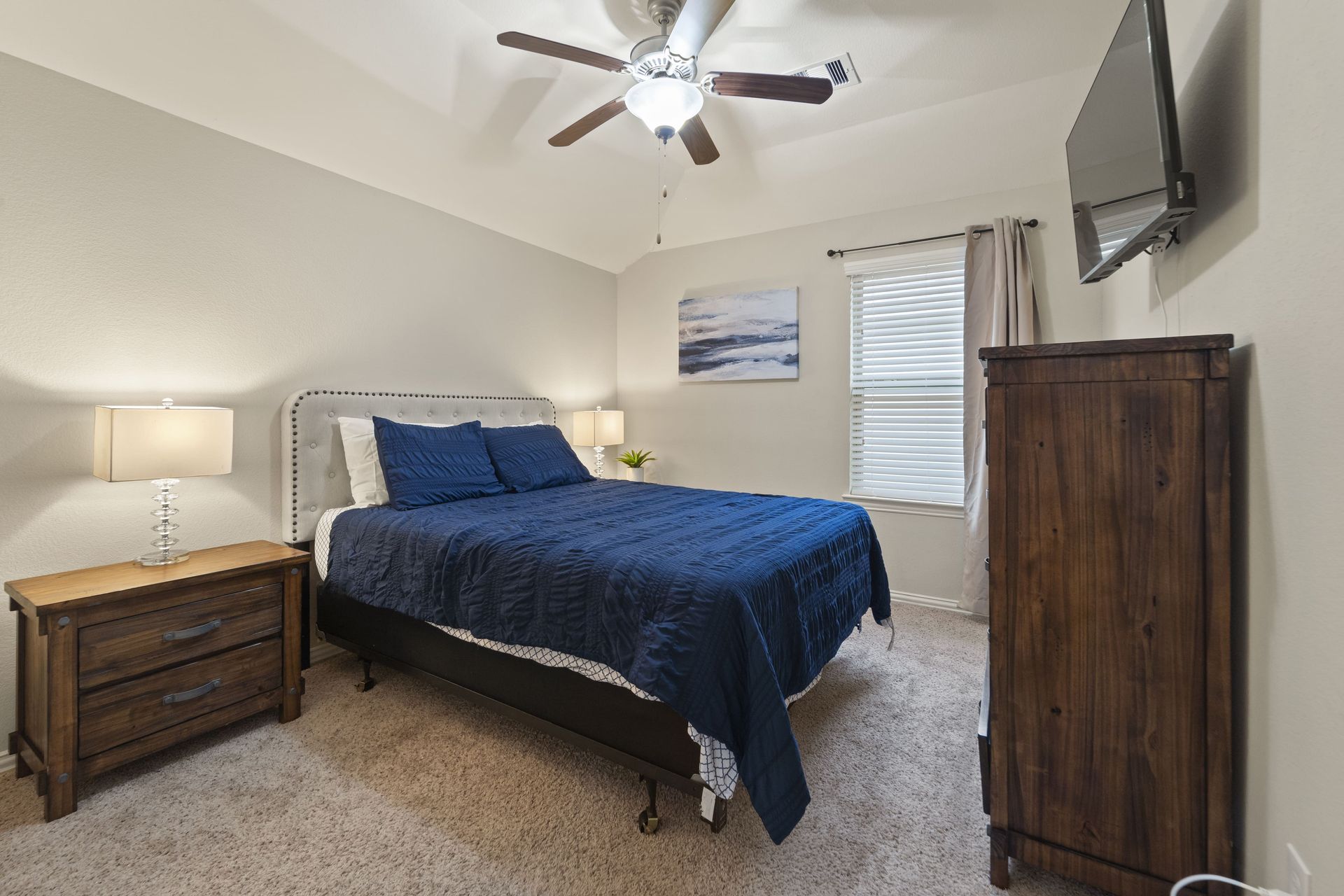 Bedroom with bed, nightstand, dresser, and TV on wall. The bed has blue bedding, and the room has neutral walls and a ceiling fan.