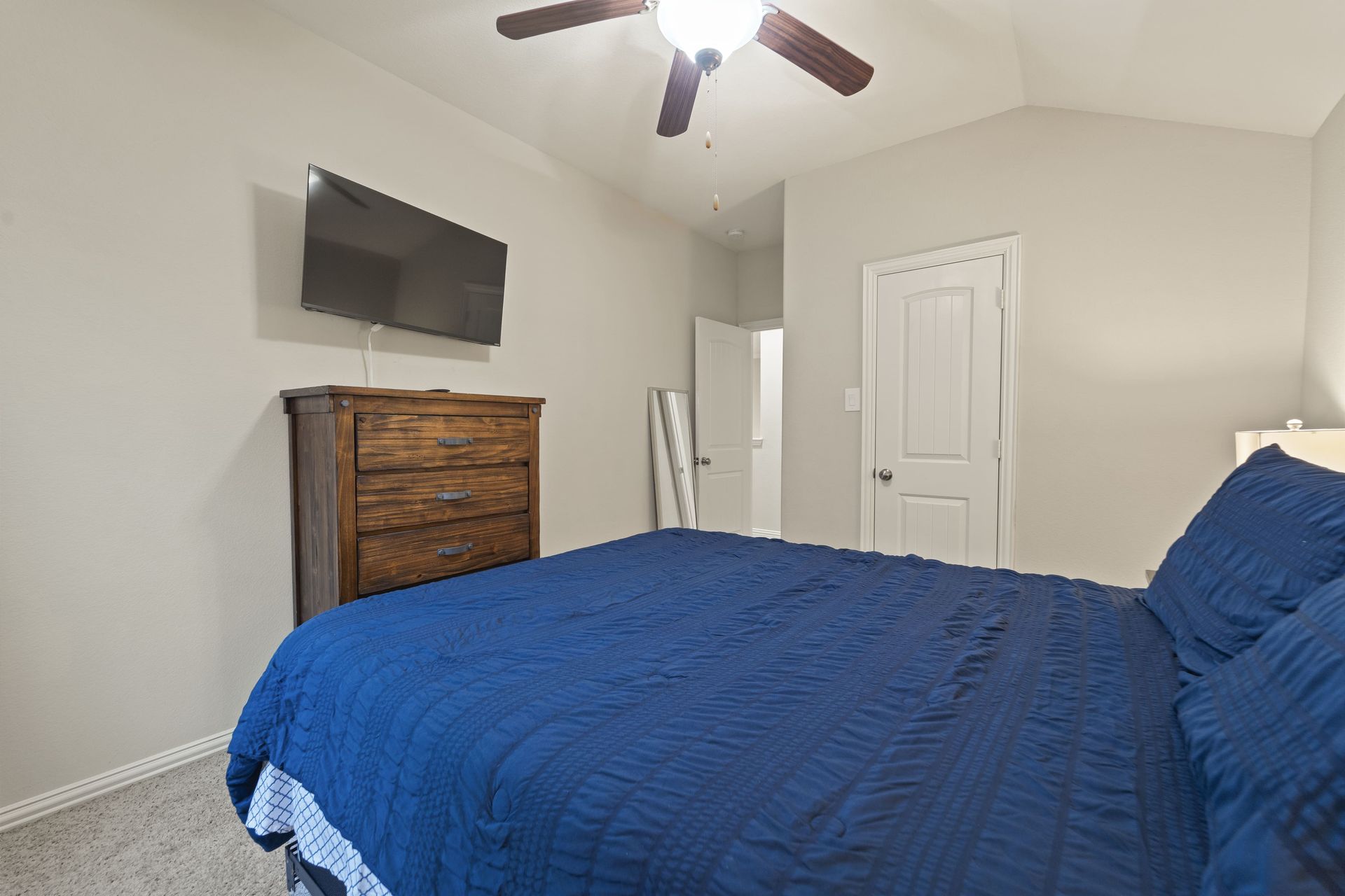 Bedroom with a bed covered in a blue comforter, a wooden dresser, and a wall-mounted TV. A door is visible in the background.
