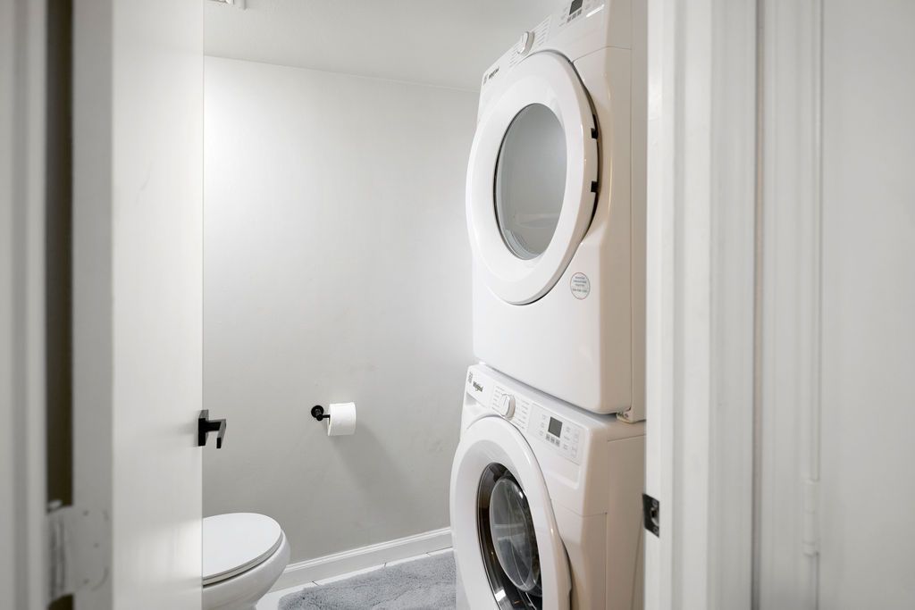 A small white bathroom with a stacked washer and dryer. A toilet sits to the left, and a roll of toilet paper is mounted on the wall.