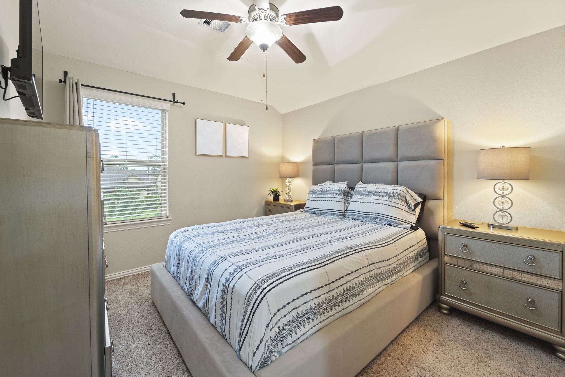 Bedroom with a bed, dressers, lamps, and a ceiling fan. Neutral tones and a window with blinds.