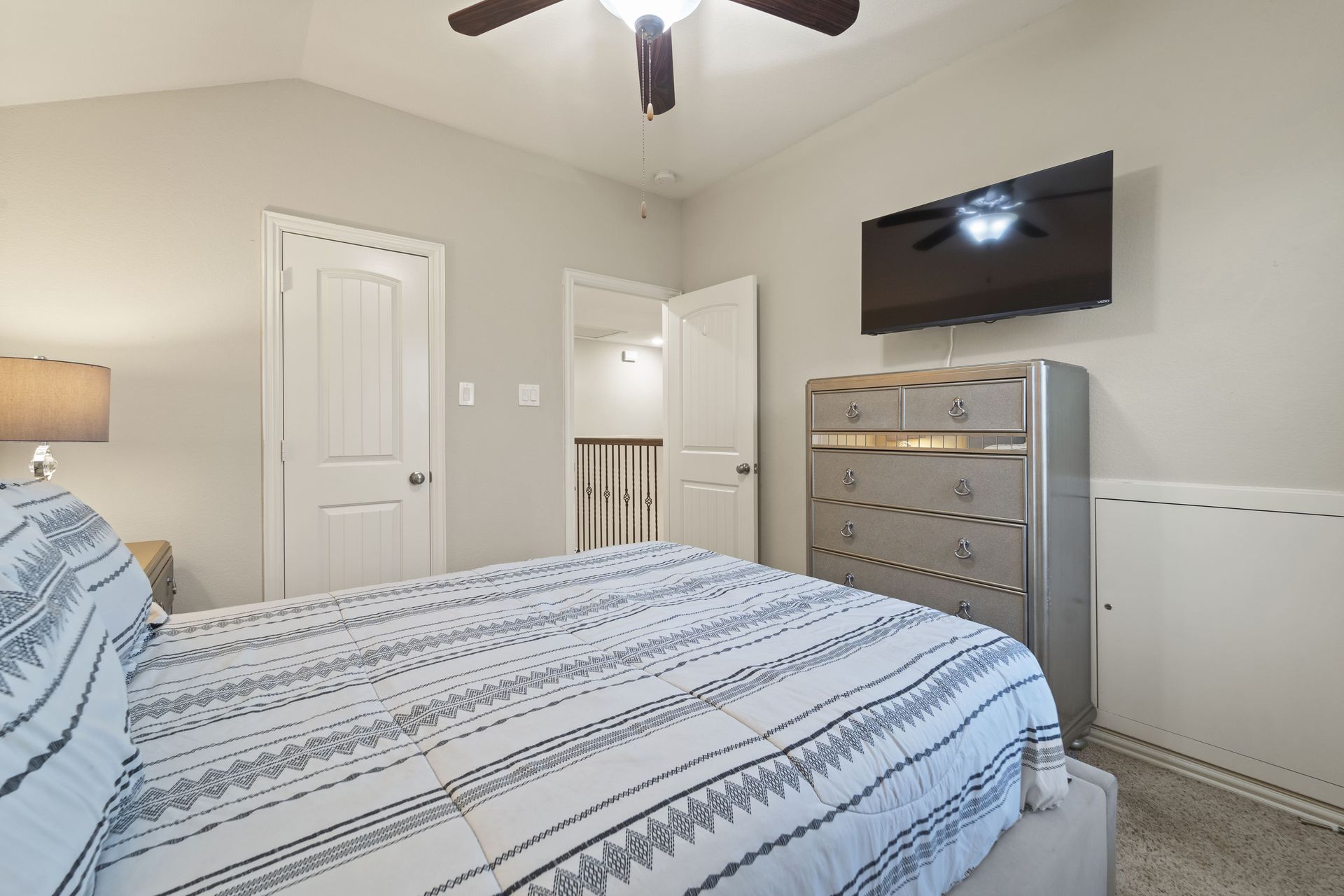 Bedroom with a bed, dresser, and a mounted TV. A door leads to a hallway. The room is painted in light neutral tones.