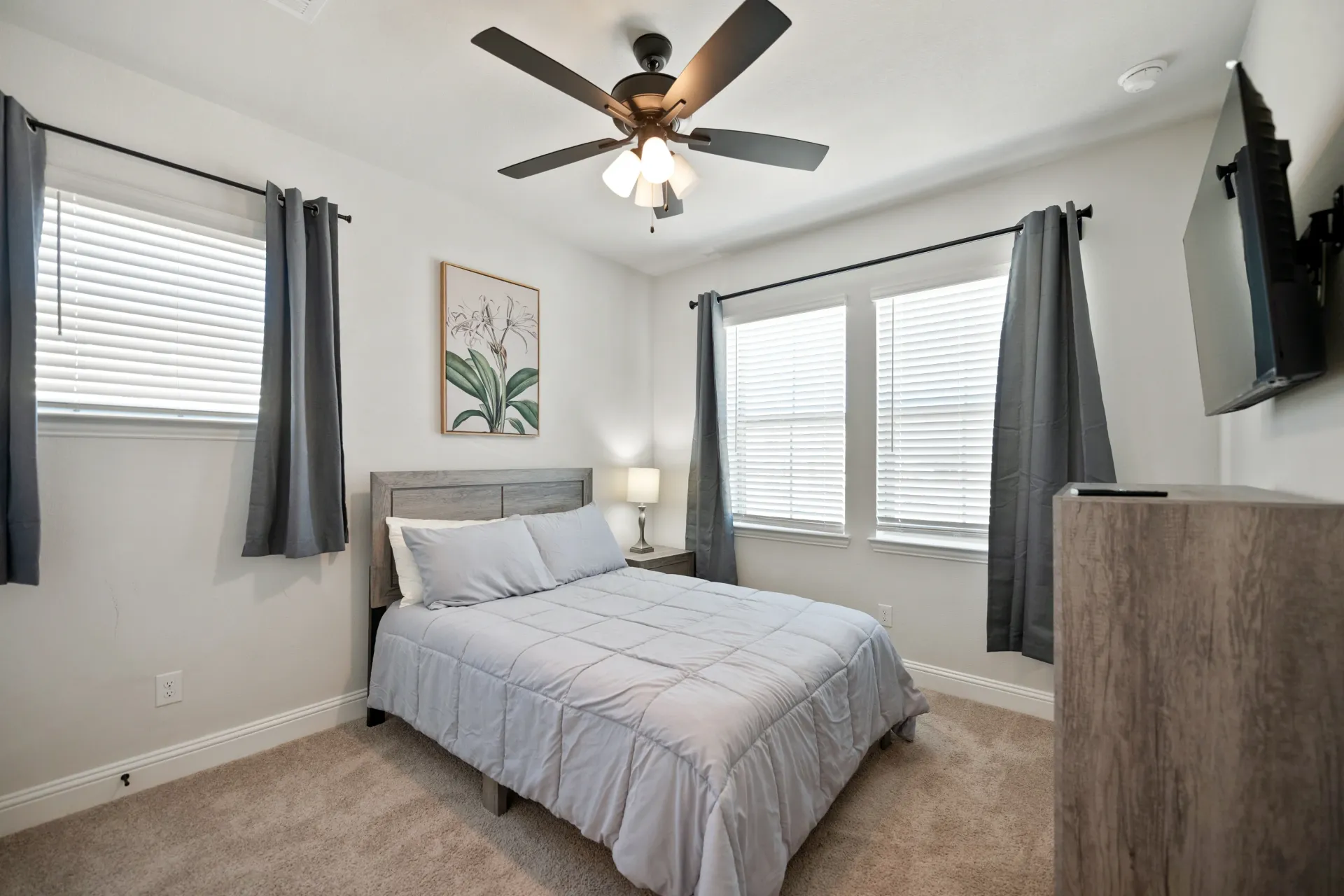 Bedroom with bed, windows, TV, and ceiling fan. Light-colored walls and carpet.