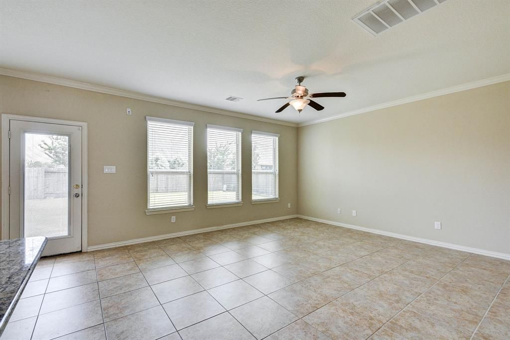 Empty, beige-walled living room with tile floors, a ceiling fan, and a door leading to a backyard. Three windows are visible.
