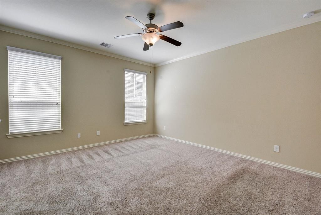 Empty bedroom with beige walls, carpet, and blinds on two windows. A ceiling fan hangs in the center.