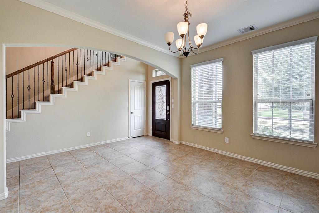 An empty living room with a staircase, arched doorway, chandelier, and two windows with blinds. Beige walls and tile floors.