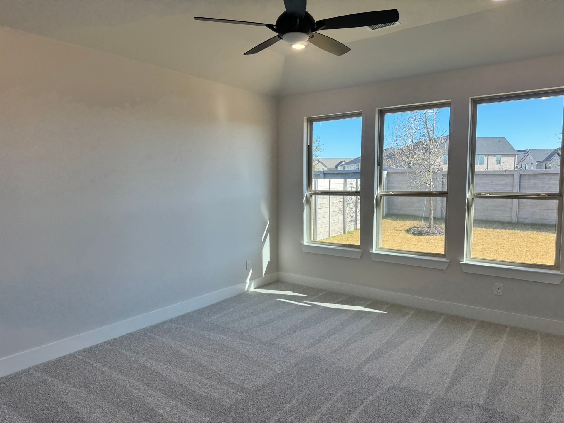 Bedroom with three windows overlooking a backyard, gray carpet, white walls, and a ceiling fan.