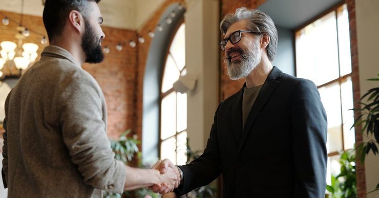 Two men shaking hands in a bright room. The younger man has a beard, the older man has gray hair and is smiling.