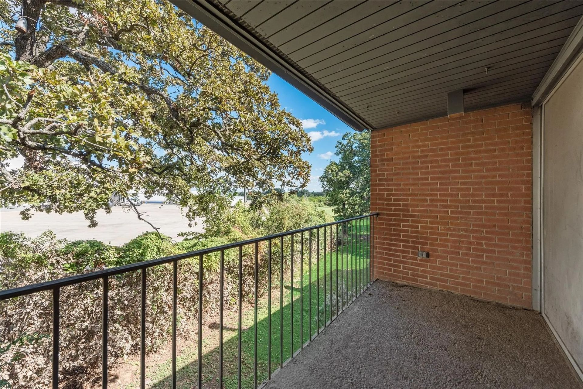 Balcony with a wrought-iron railing and a red brick wall, overlooking greenery and a road under a bright blue sky.