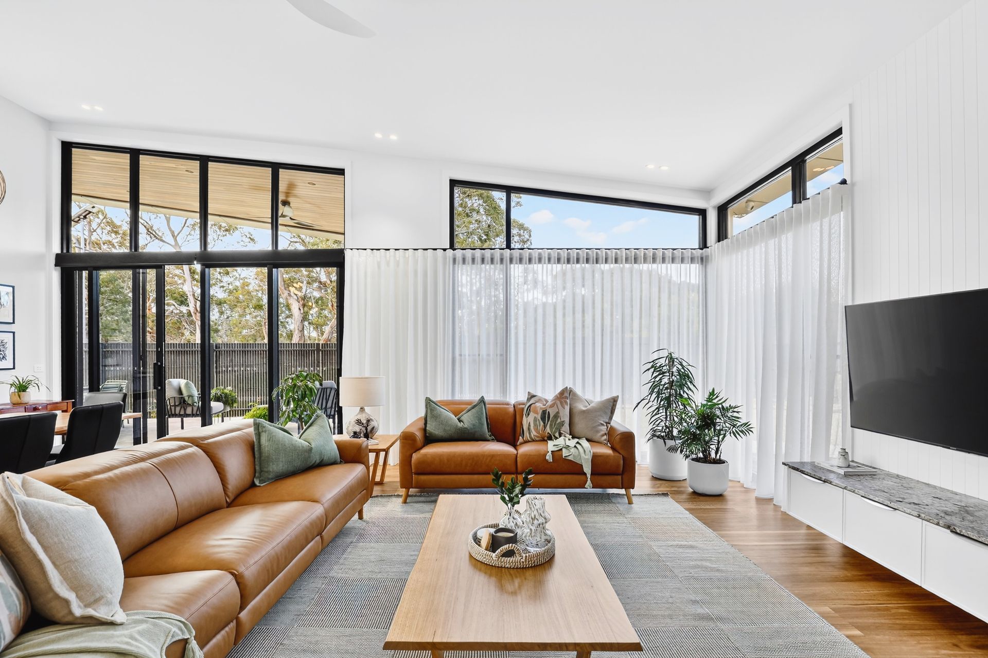 Living room with tan leather sofa, large windows, white curtains, and TV.