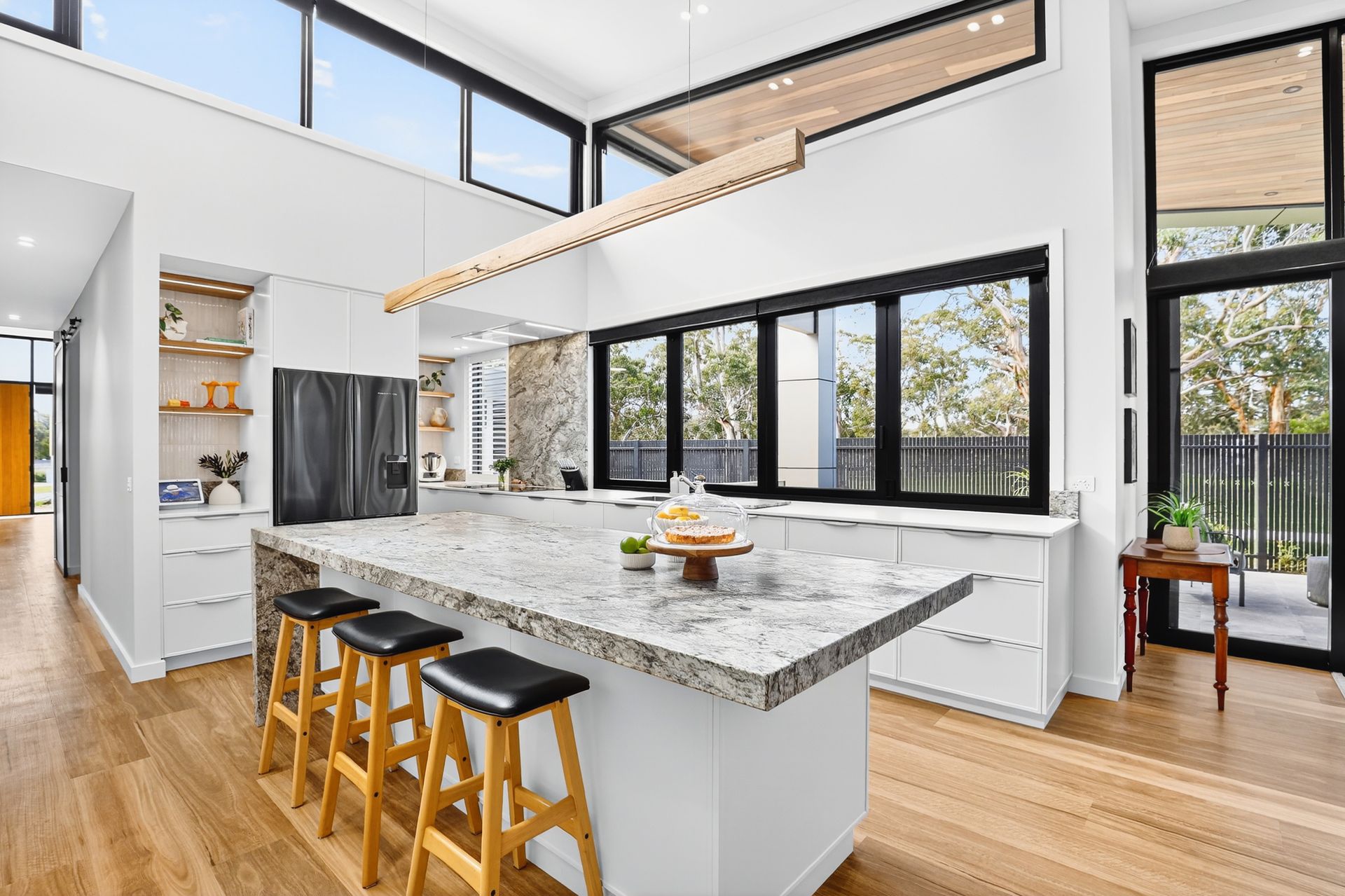 Modern kitchen with large island, granite countertop, stools, wood floor, and black-framed windows, and double-height ceiling.