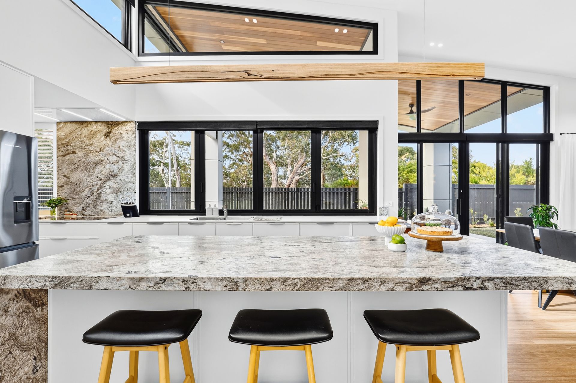 Modern kitchen with granite countertops, black bar stools, and a large window overlooking trees.