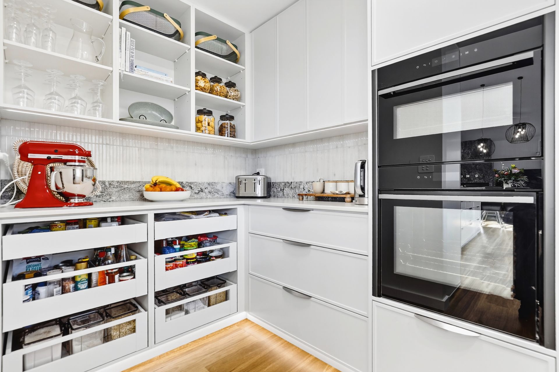 Modern, white pantry with pull-out drawers, open shelving, red mixer, built-in oven, and countertop with snacks.