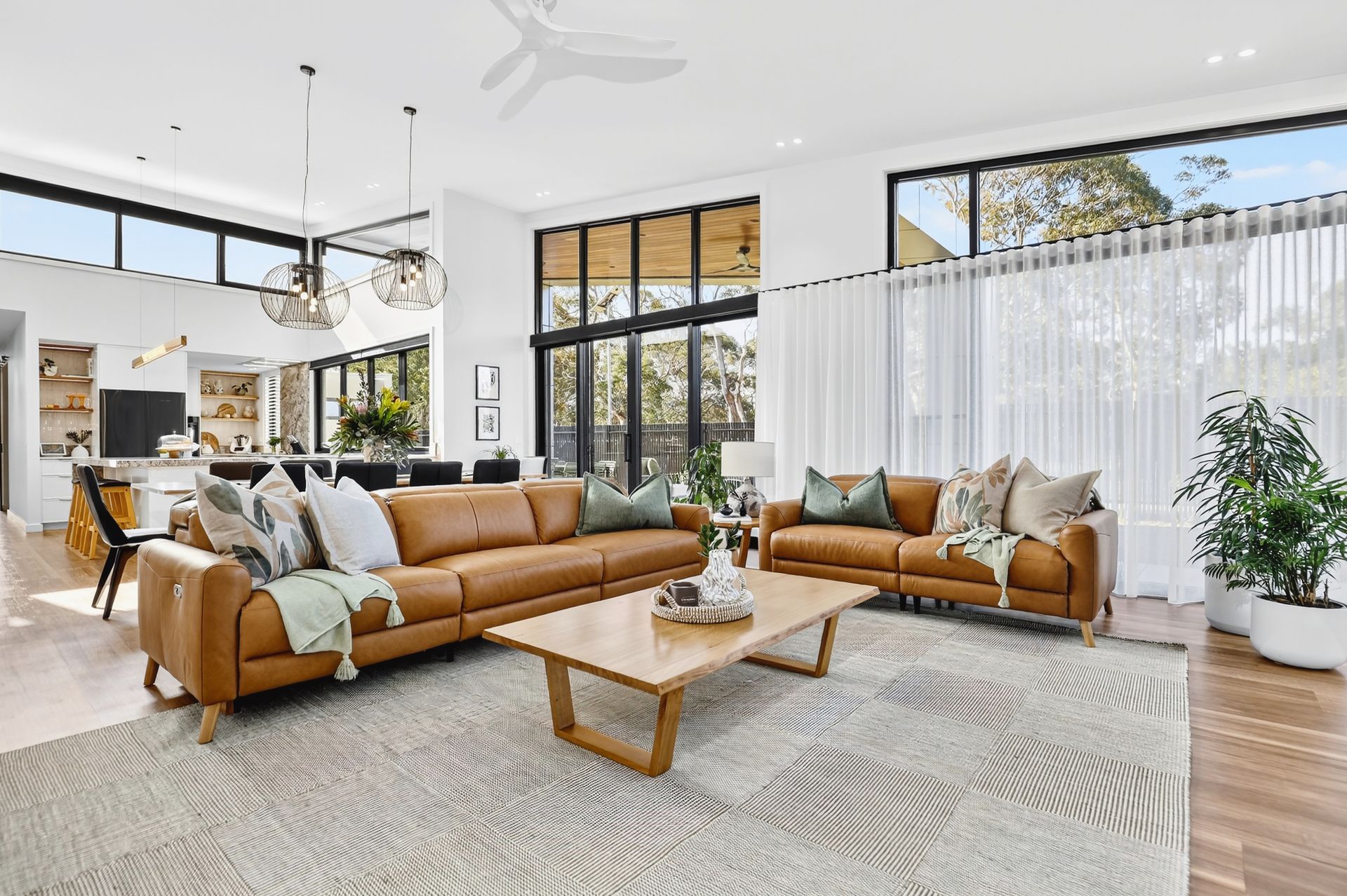 Spacious living room with brown leather sofas, wooden coffee table, and large windows overlooking a yard.