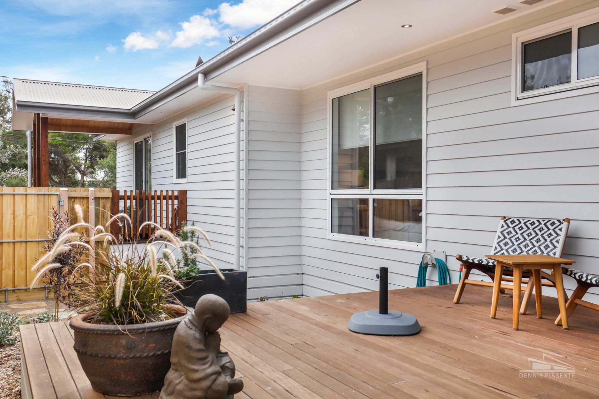 Outdoor deck with wooden planks, white house siding, seating area, and decorative plants and a statue.