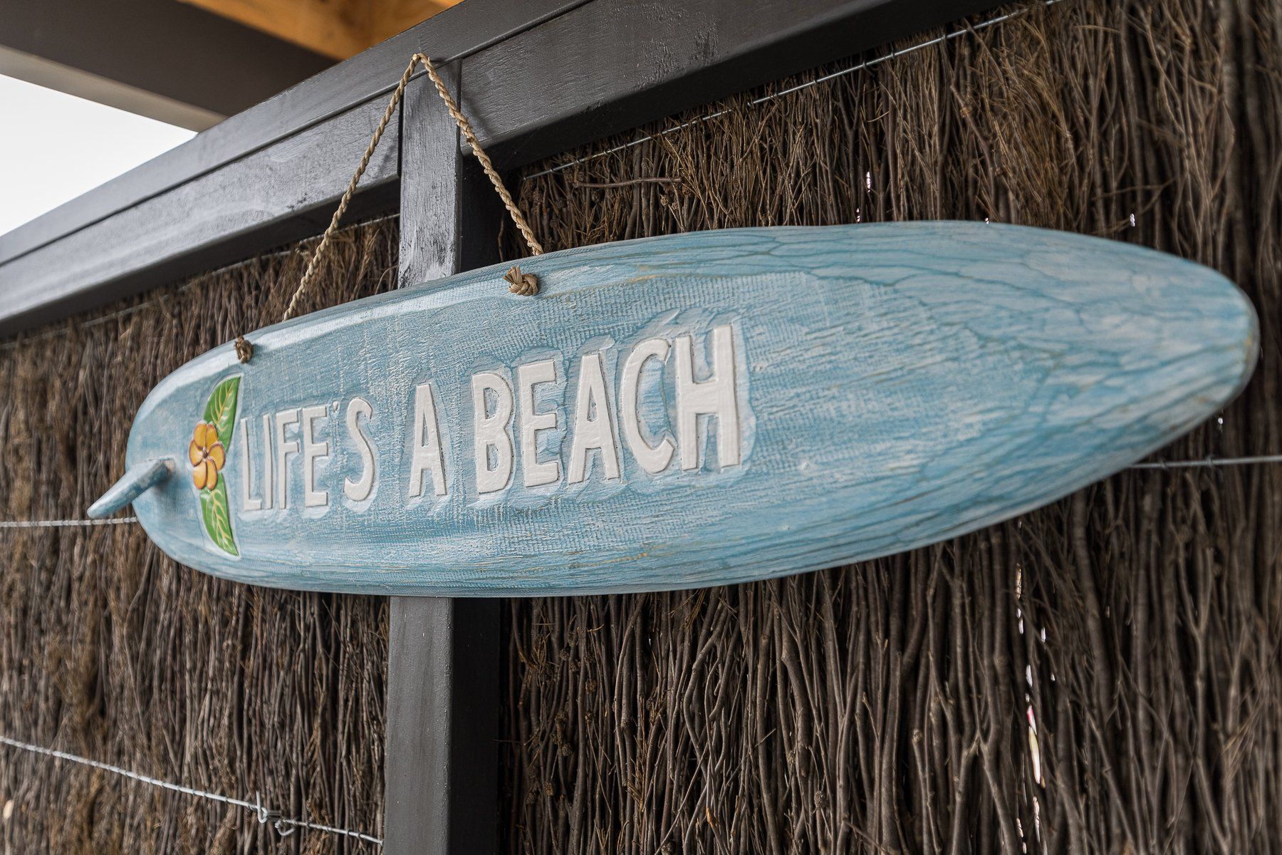 A blue surfboard with the words `` life 's a beach '' hanging on a fence.