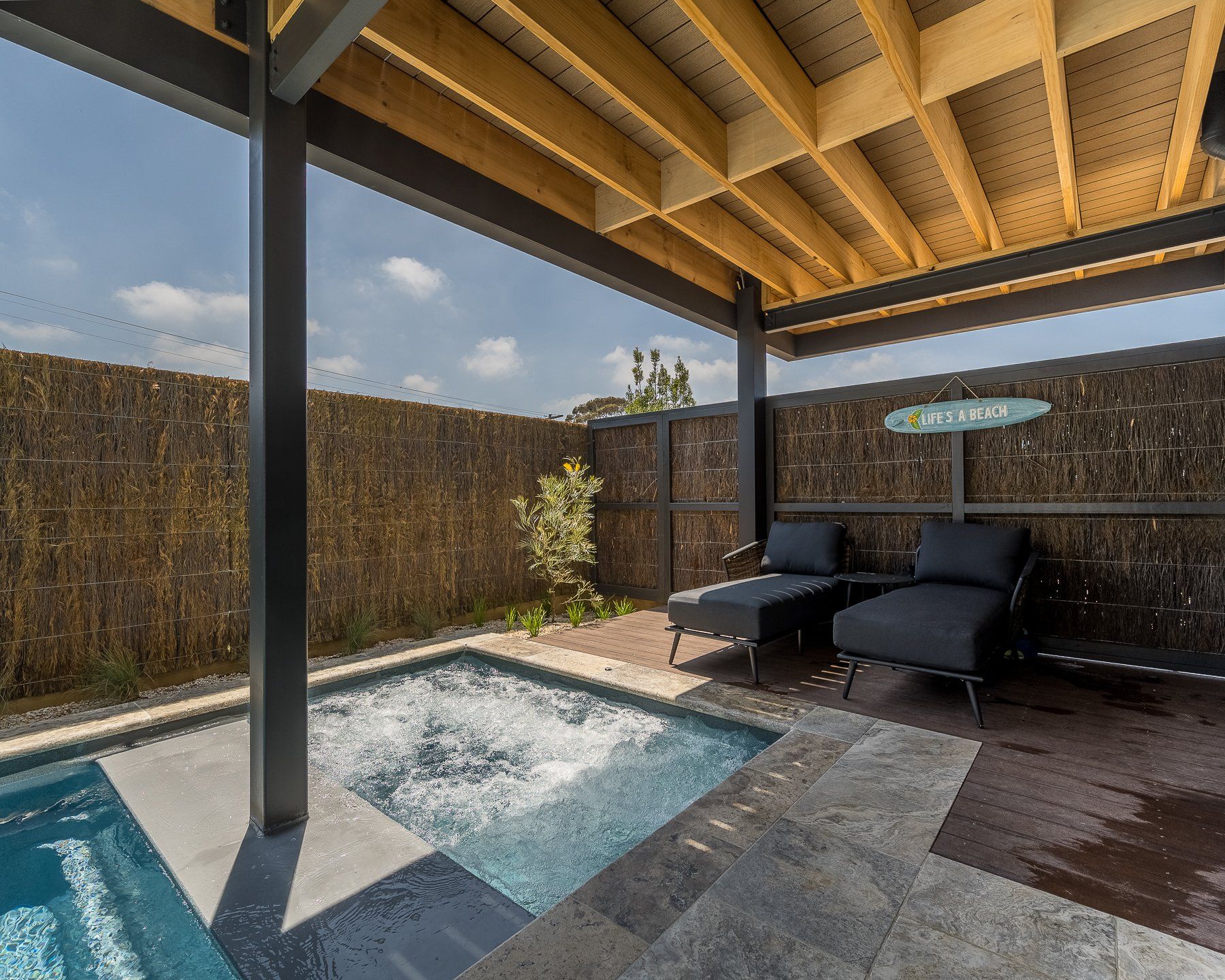 A small swimming pool is under a covered area with chairs and a surfboard.