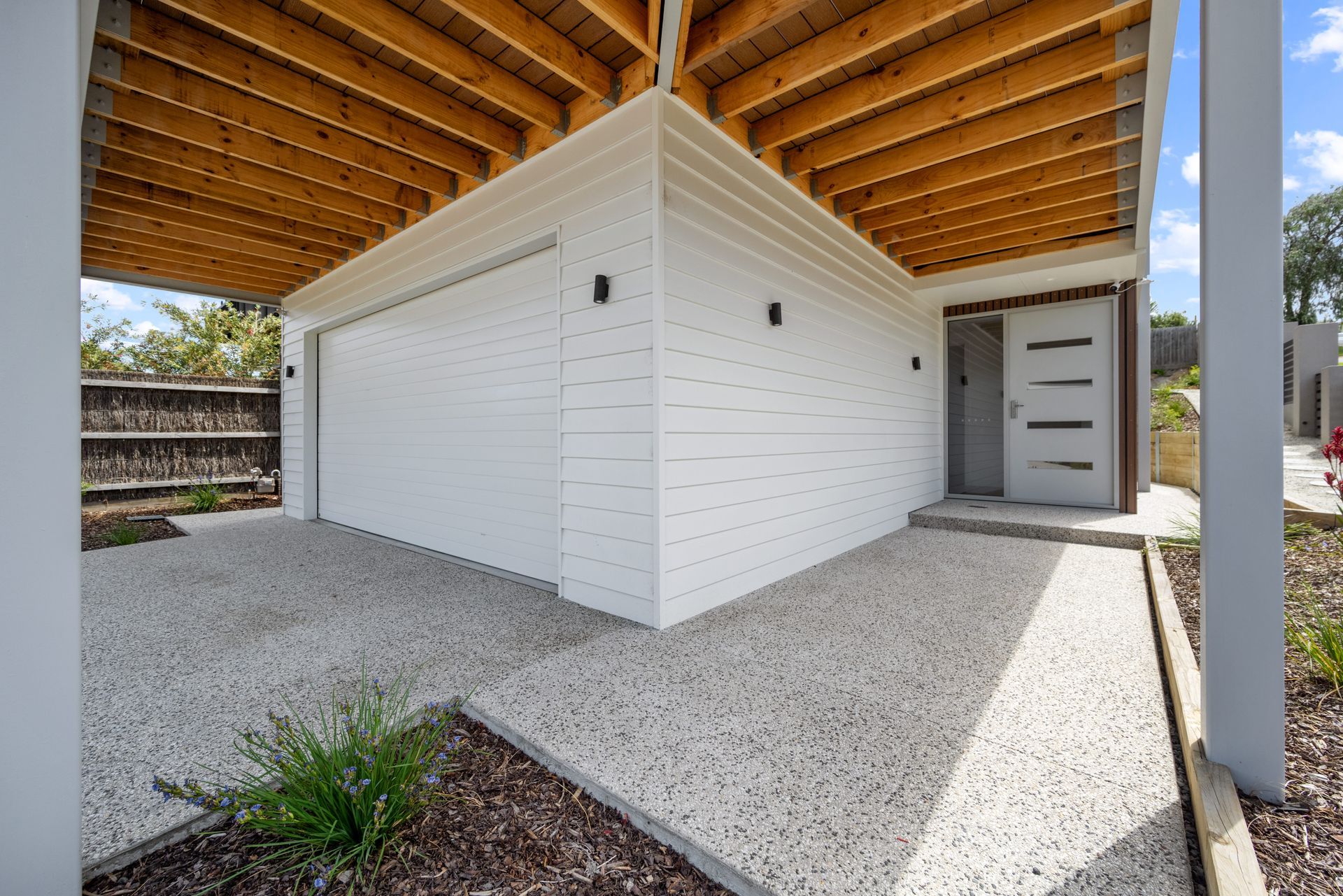 Exterior view of a white modern home with a garage and front entrance. Gravel driveway, wooden ceiling, and a shrub are visible.