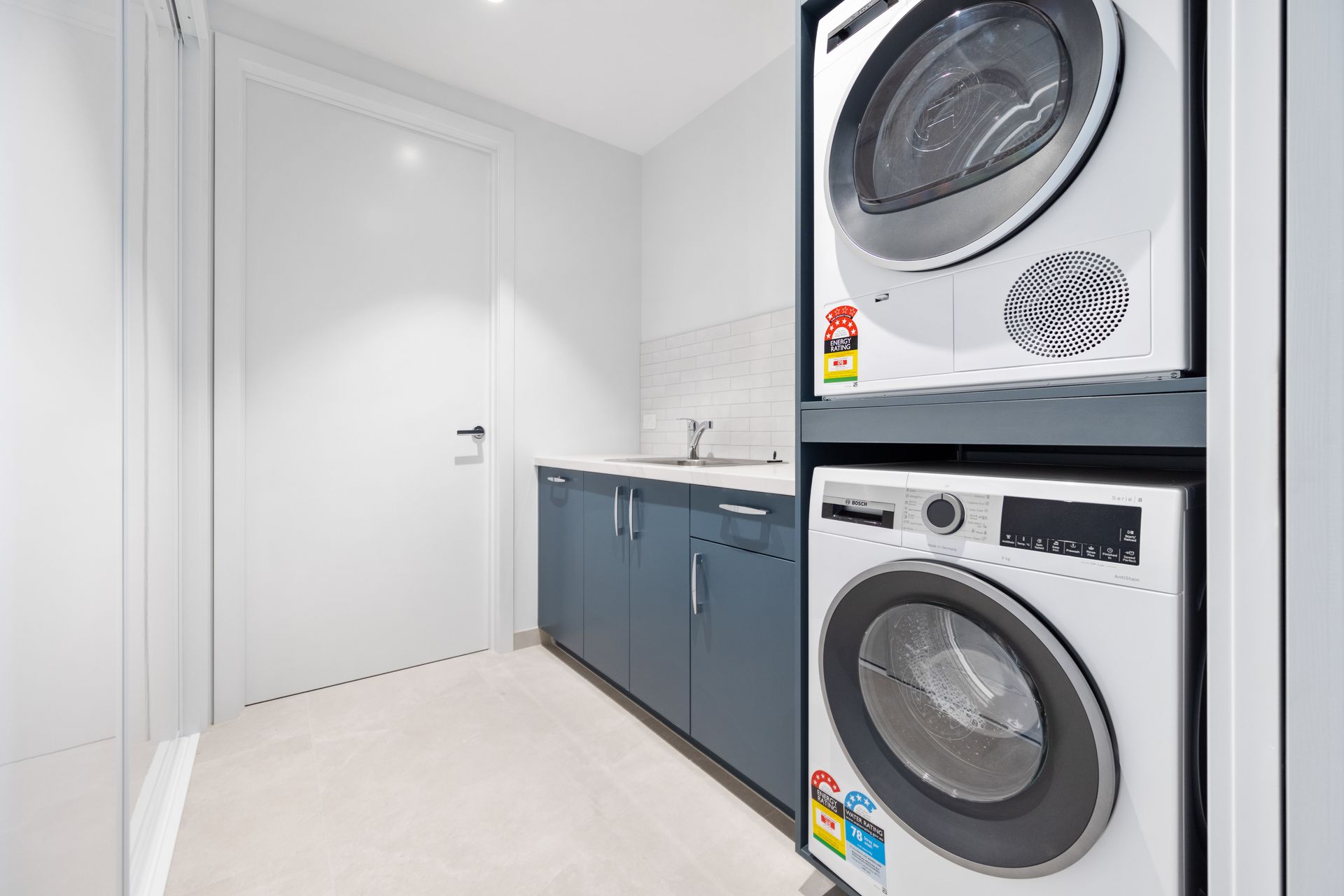 Laundry room with a stacked washer and dryer, blue cabinets, and a small sink. The room is mostly white.