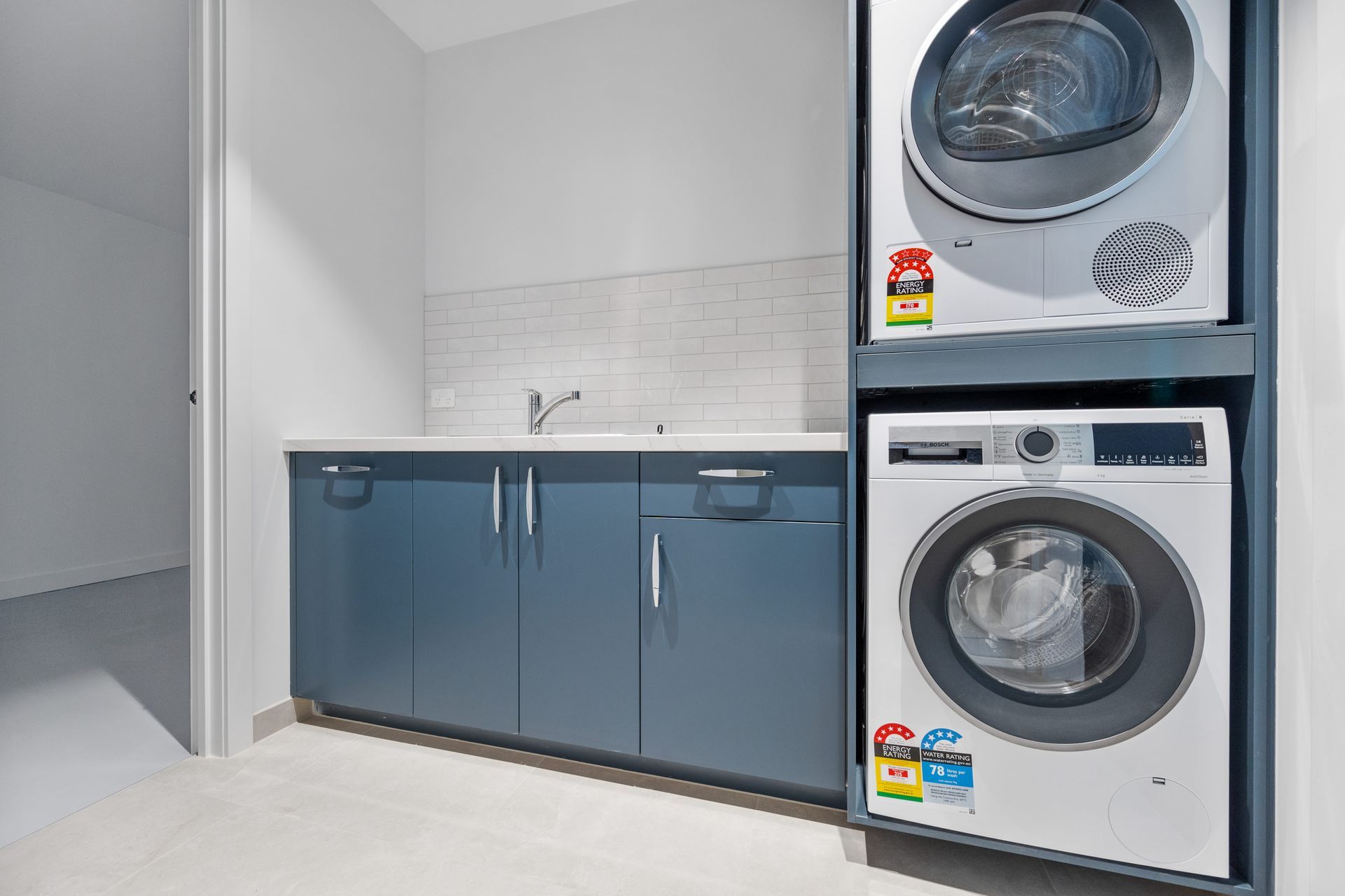 Laundry room with stacked washer and dryer, blue cabinets, a sink, and a white tiled backsplash.