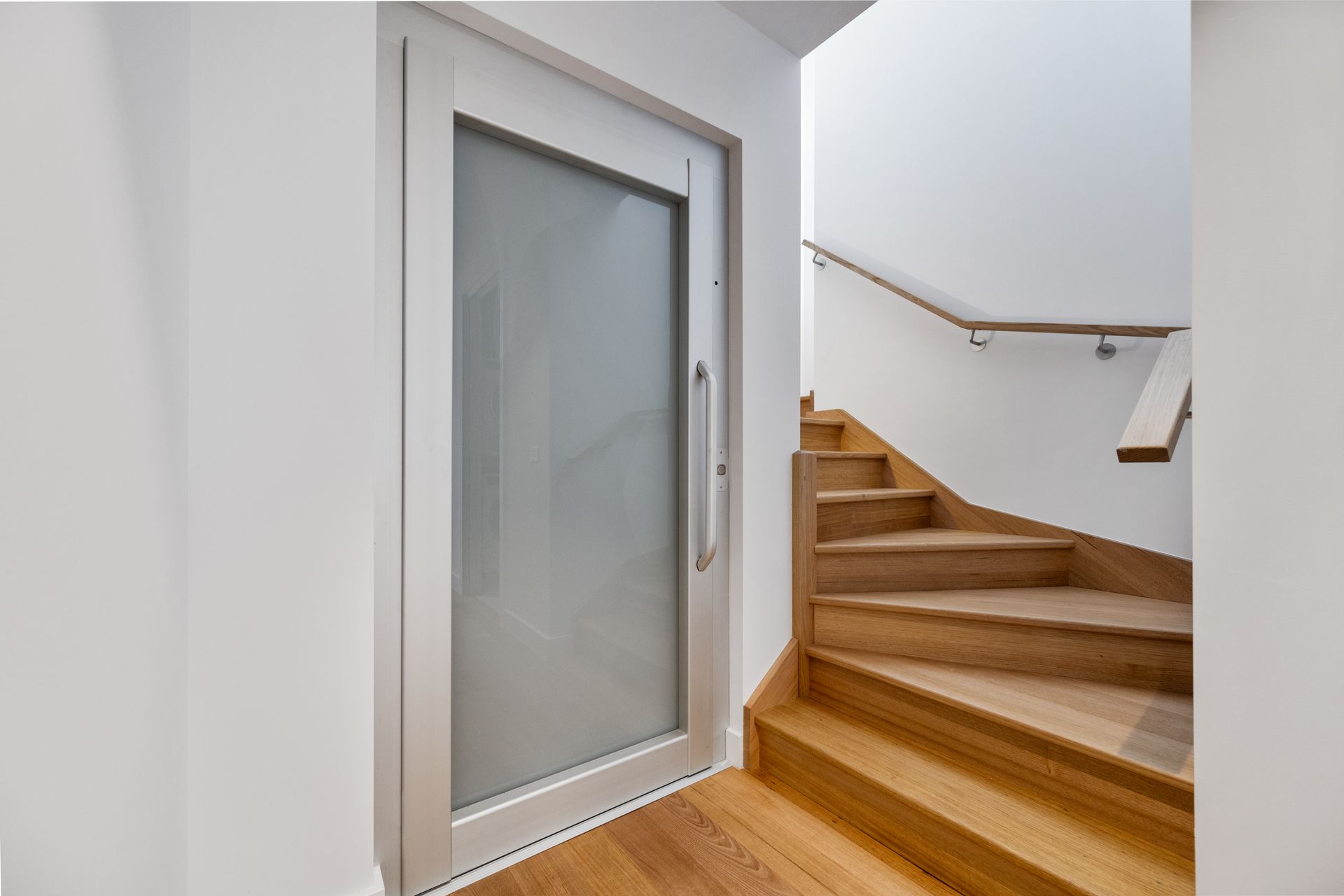 Door with frosted glass panel next to a wooden staircase with a handrail. The walls are white, and the floor is wood.