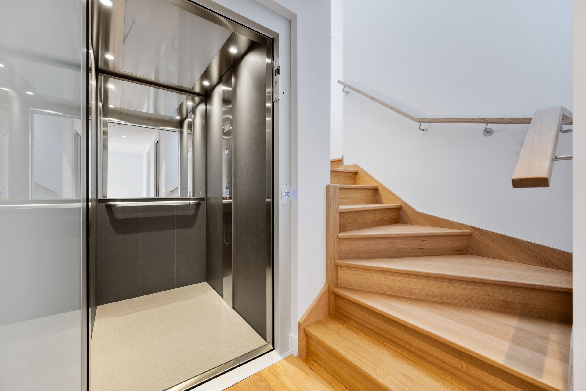 An open elevator next to a wooden staircase. The elevator has chrome doors and a beige floor. The staircase has wooden steps and a white wall.