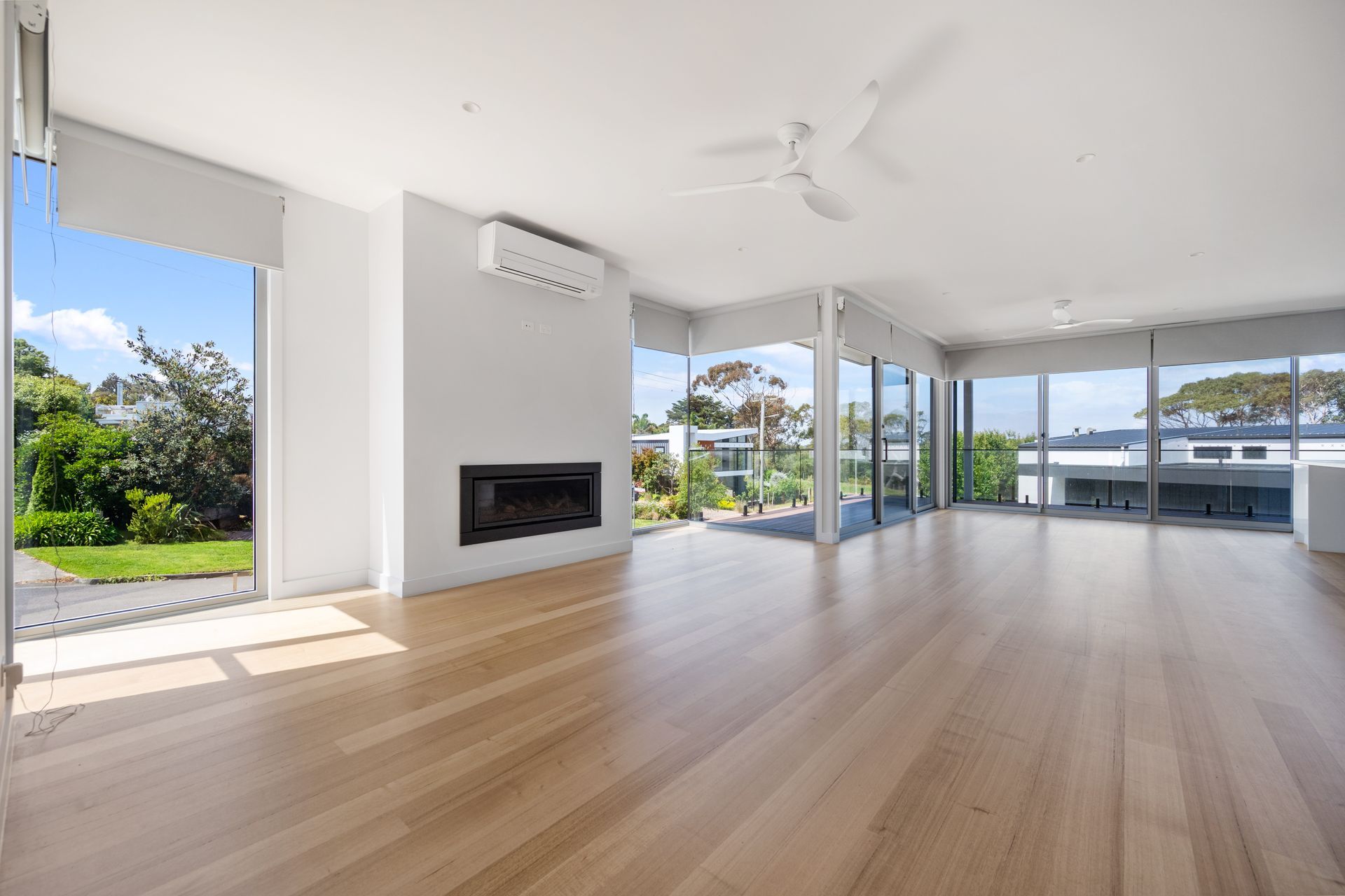 Bright, modern living room with large windows overlooking greenery and a partial view of a building. Features hardwood floors, a fireplace, and air conditioning unit.