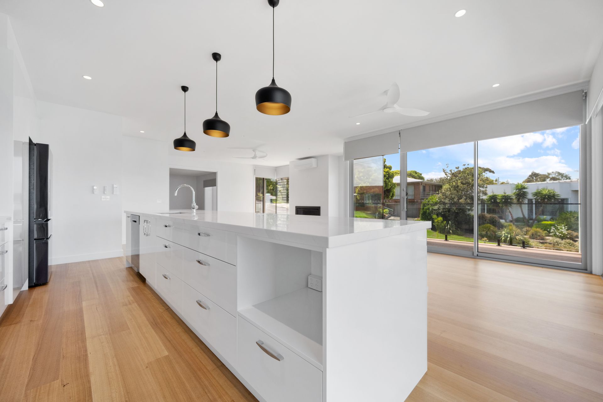 Spacious, modern kitchen with a large white island, wood floors, and floor-to-ceiling windows overlooking a sunny outdoor scene.