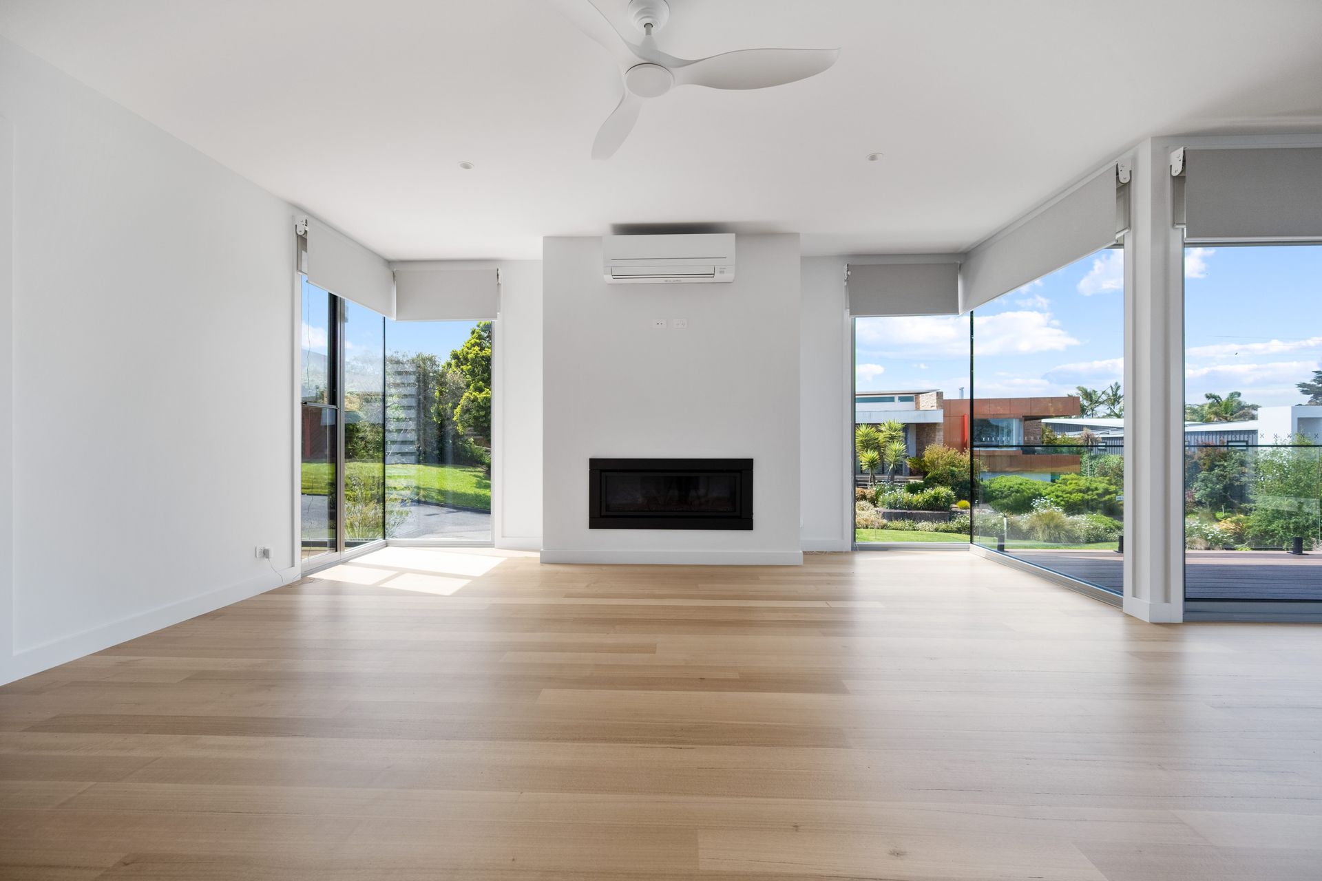 Empty living room with wood floors, large windows, fireplace, and a ceiling fan. Sunlight streams in.