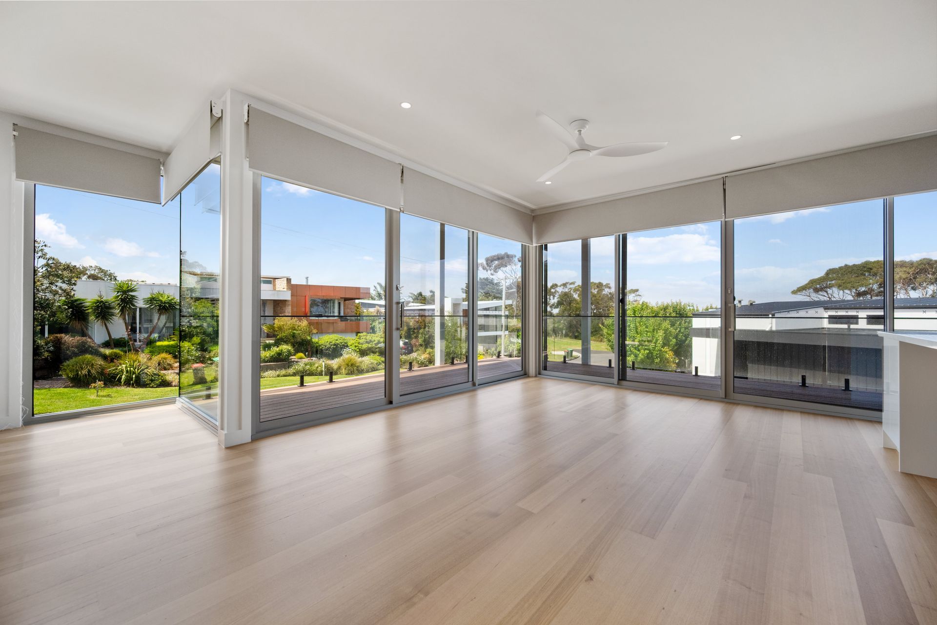 Bright, empty room with floor-to-ceiling windows overlooking a green yard. The room features light wooden floors and a white ceiling.