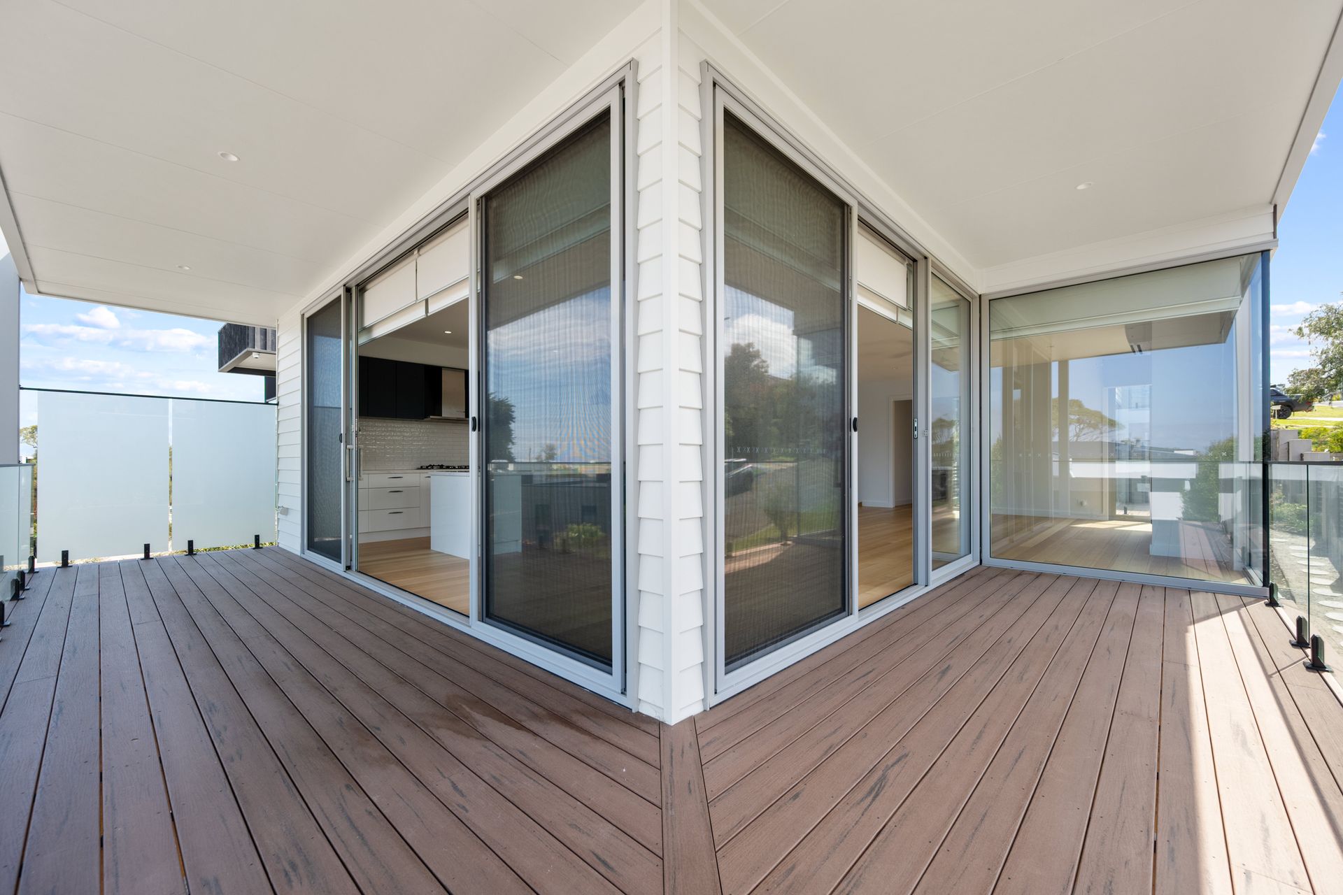 A modern deck with brown wood-like flooring leads to a bright interior with large sliding glass doors.