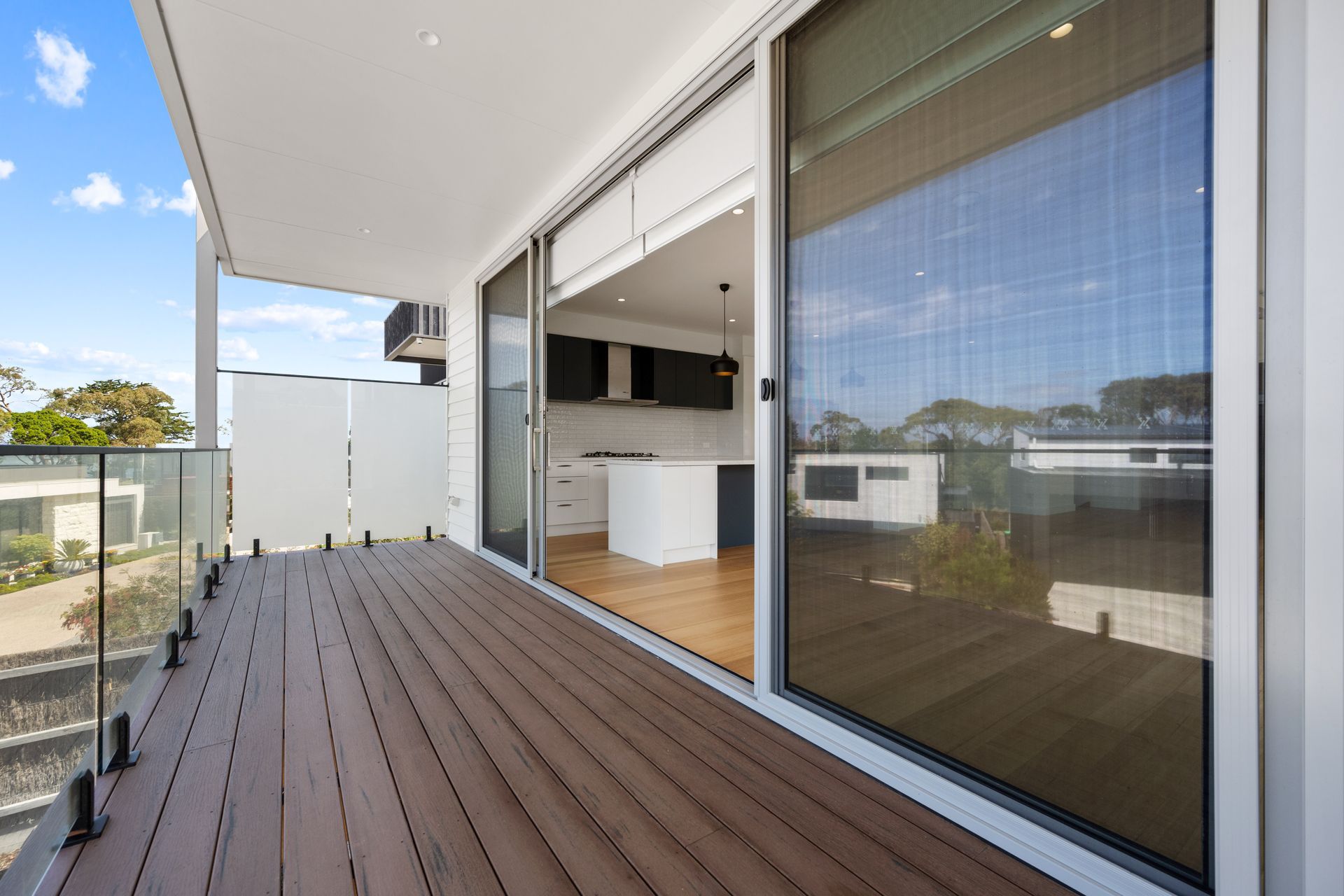 Balcony with brown deck and glass railing, leading into a modern kitchen with large sliding glass doors.