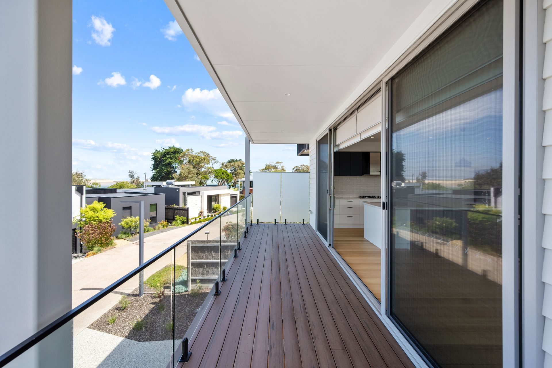 Balcony with composite decking, glass railings, and sliding glass door looking toward a modern neighborhood on a sunny day.