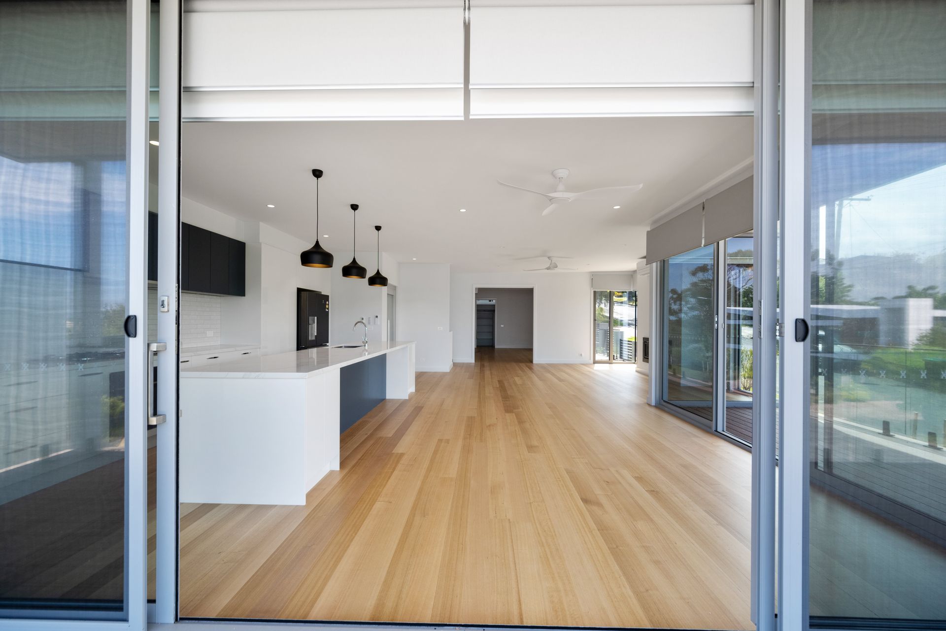 View from open sliding doors into a bright, modern living space with wood floors, a white kitchen, and large windows.