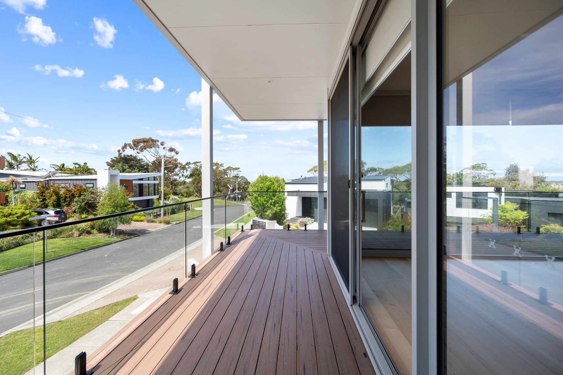 Balcony with composite decking, glass railings, and sliding glass door overlooking a residential street on a sunny day.