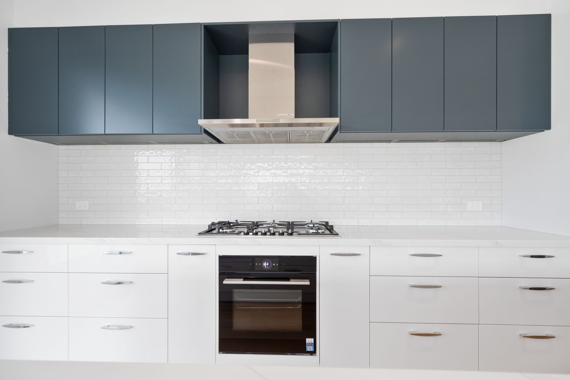 Modern kitchen with white countertops, white subway tile backsplash, and a stainless steel range hood. Upper cabinets are blue-gray, and lower cabinets are white.