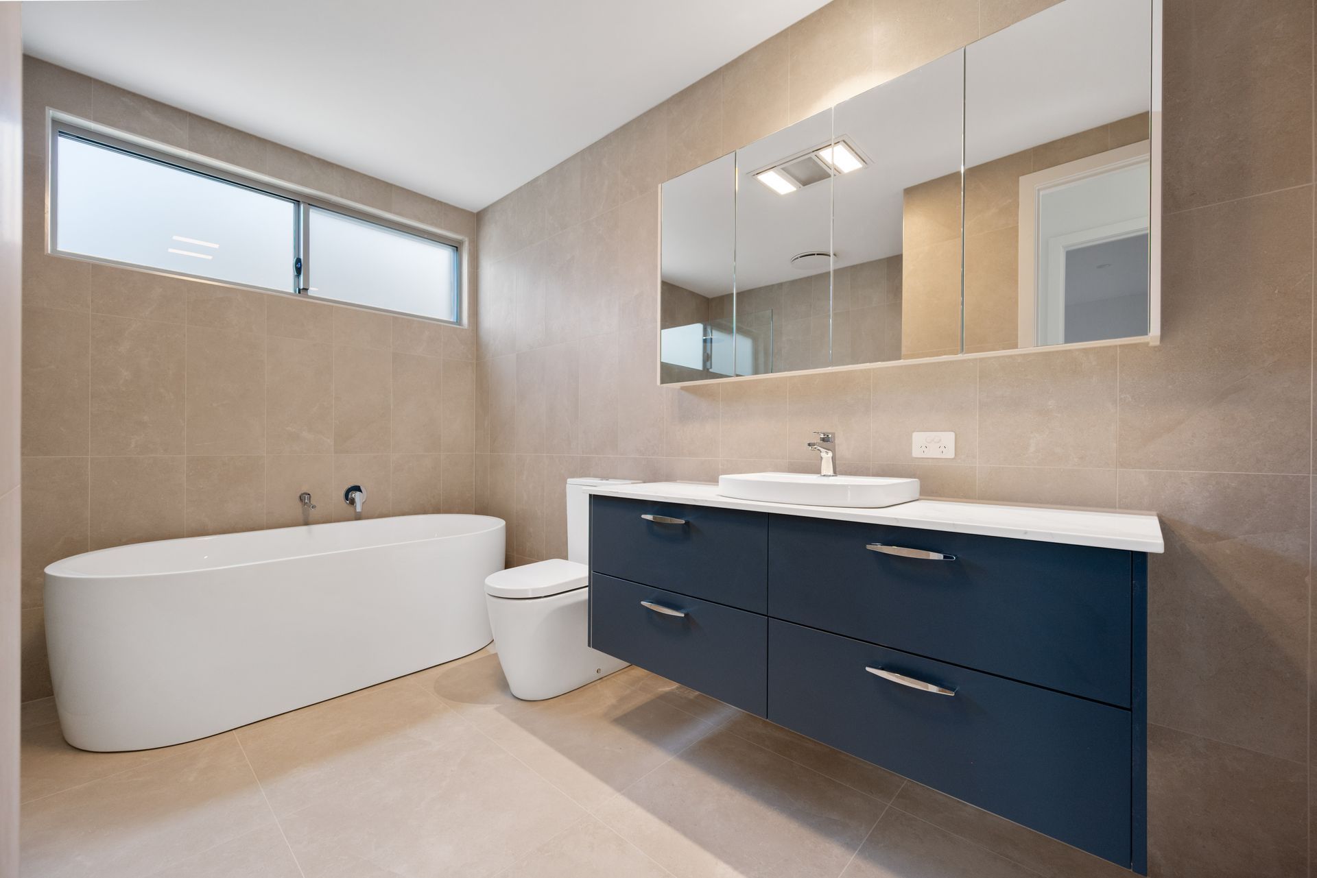 Modern bathroom with a blue vanity, white sink and tub, and beige tiles.