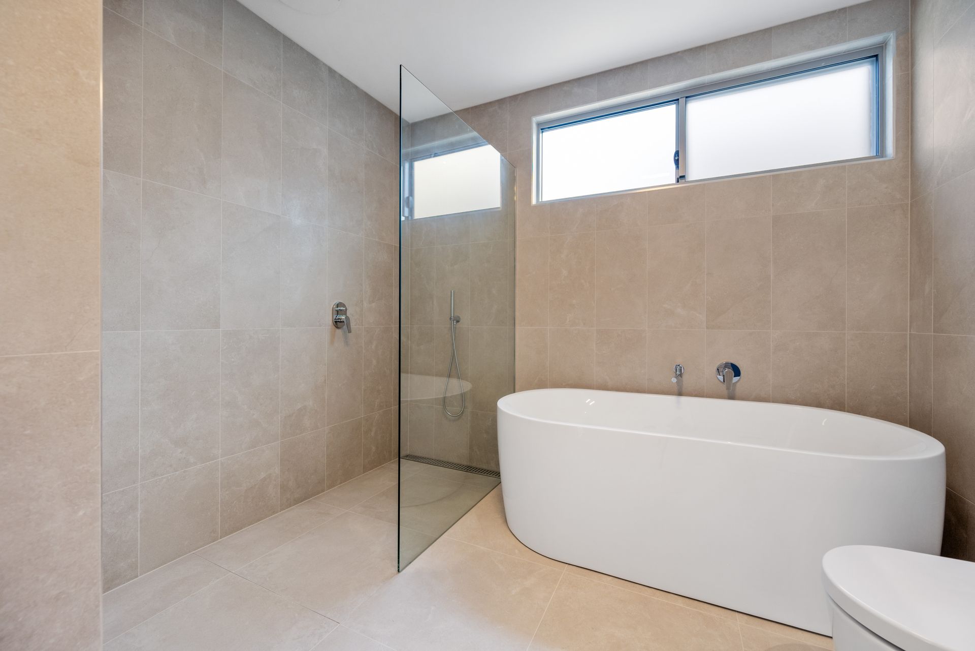 Modern bathroom with a white oval tub, glass shower, and neutral-colored tiled walls. Natural light streams in from a window.