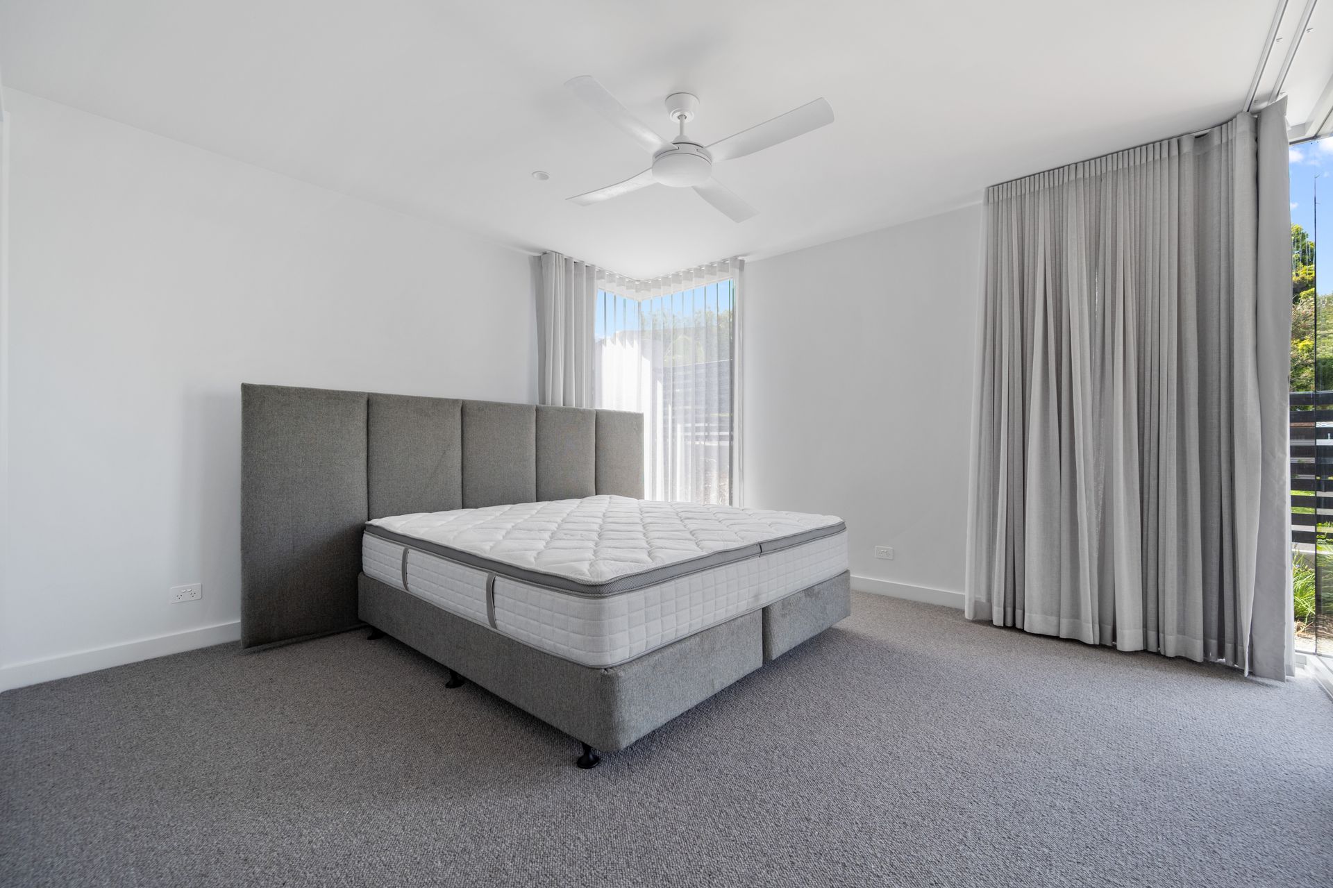 Bedroom with a large gray upholstered bed, headboard, and gray carpet. White walls and ceiling with a ceiling fan, light gray curtains, and windows.